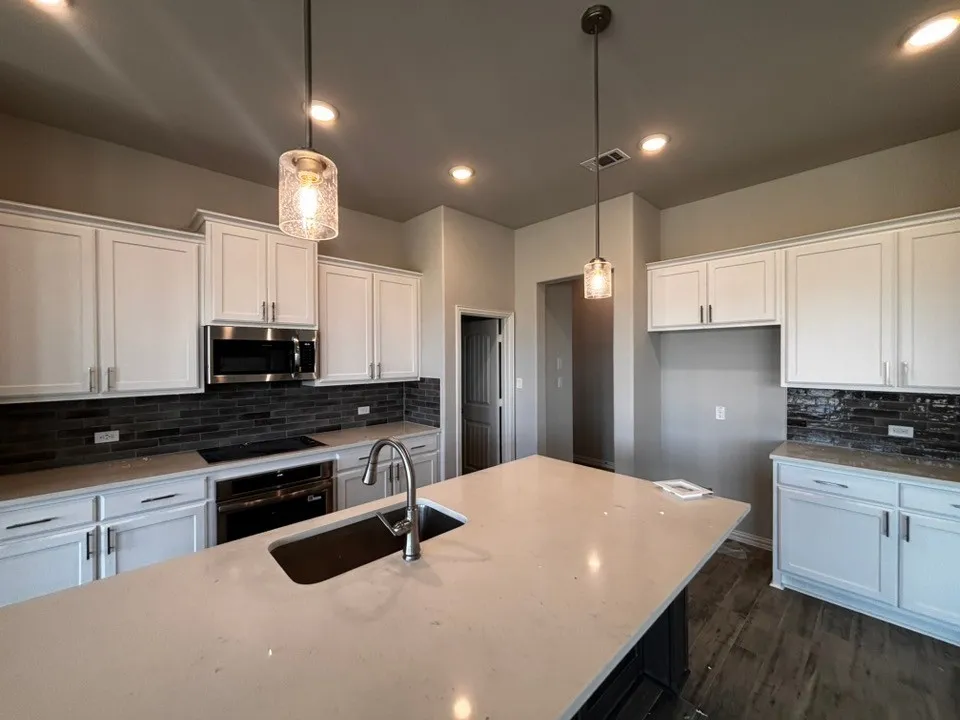 White shaker-style cabinetry with chrome hardware, dark subway tile backsplash, stainless steel built-in microwave, integrated cooktop, and an island with an undermount sink and gooseneck faucet
