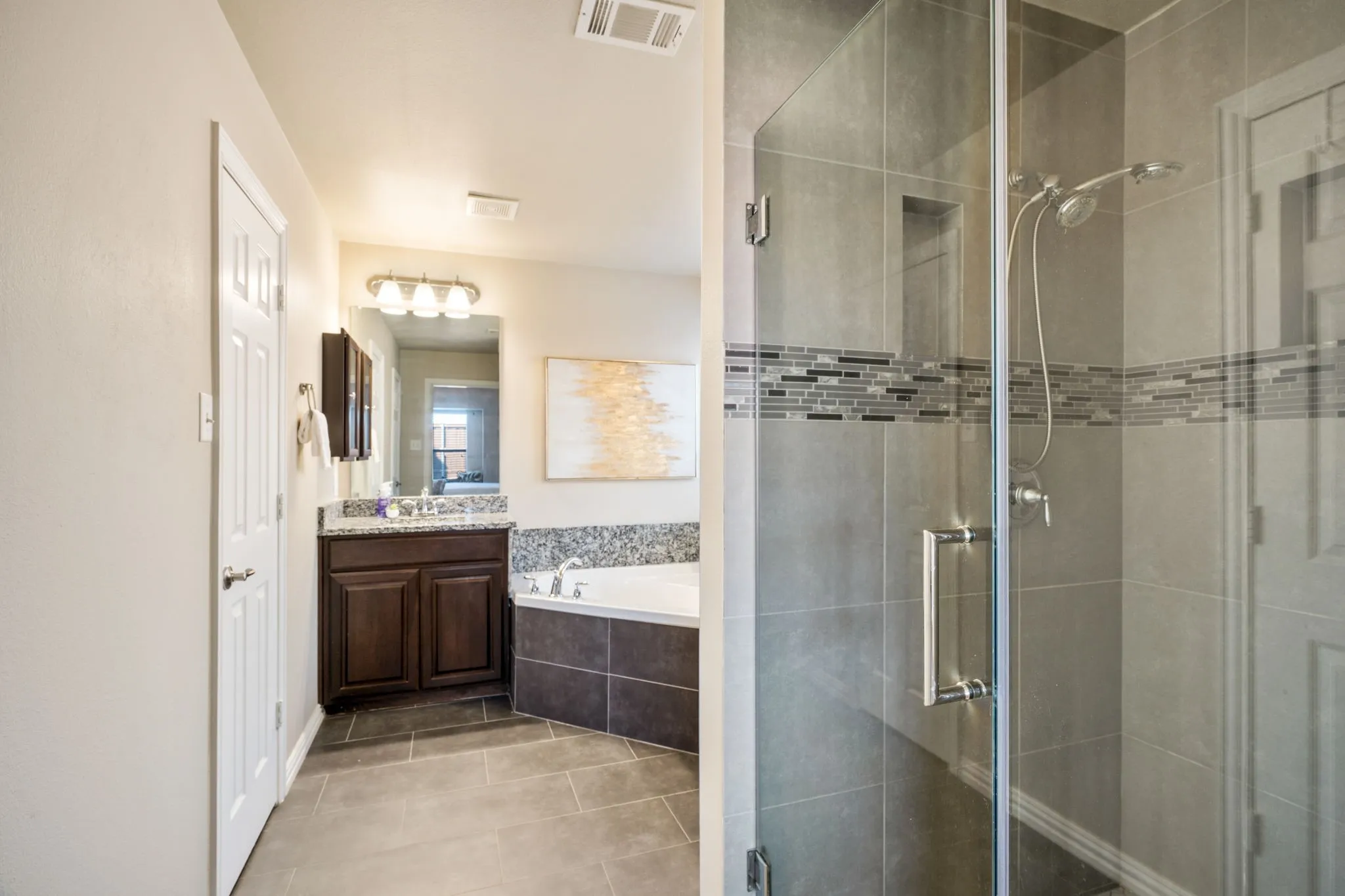 Enclosed shower with tiled walls and glass door, integrated built-in niche, corner soaking tub, vanity with a dark wood-finish cabinet and stone-finish countertop, and tile flooring