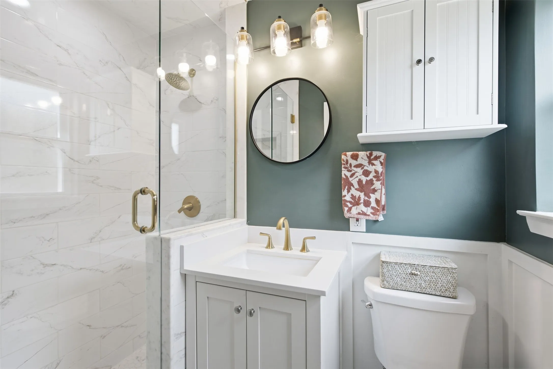 Glass-enclosed shower with subway tile surround, white vanity with integrated sink, gold-toned fixtures, and a wall-mounted cabinet