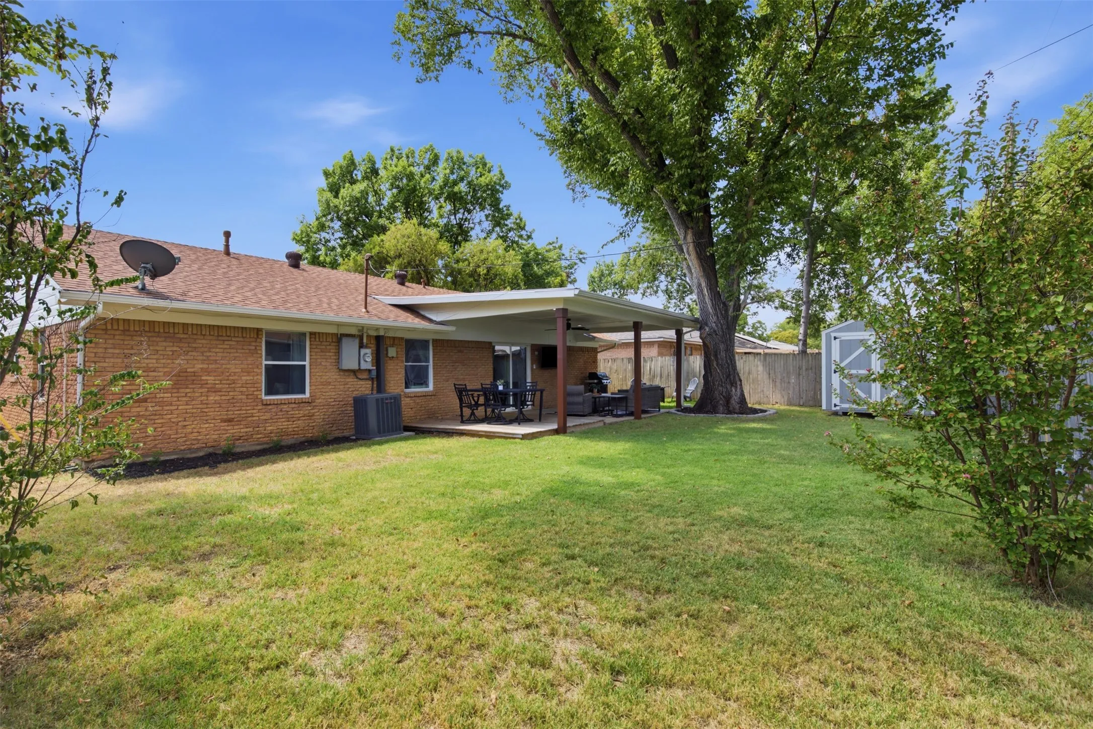 Expansive backyard featuring a brick exterior, covered patio, mature tree, and a storage shed