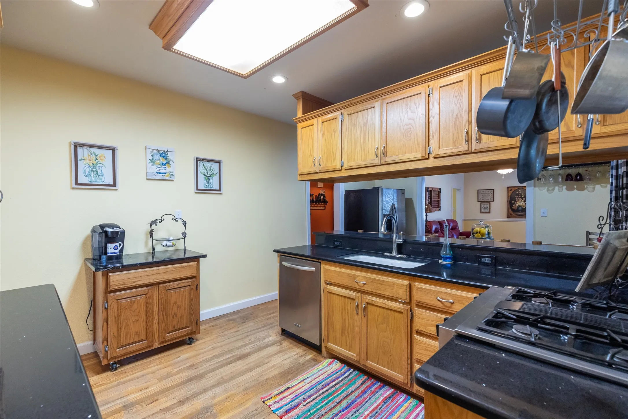 Recessed lighting, wood-finish flooring, wood cabinetry, black countertops, and a stainless steel dishwasher