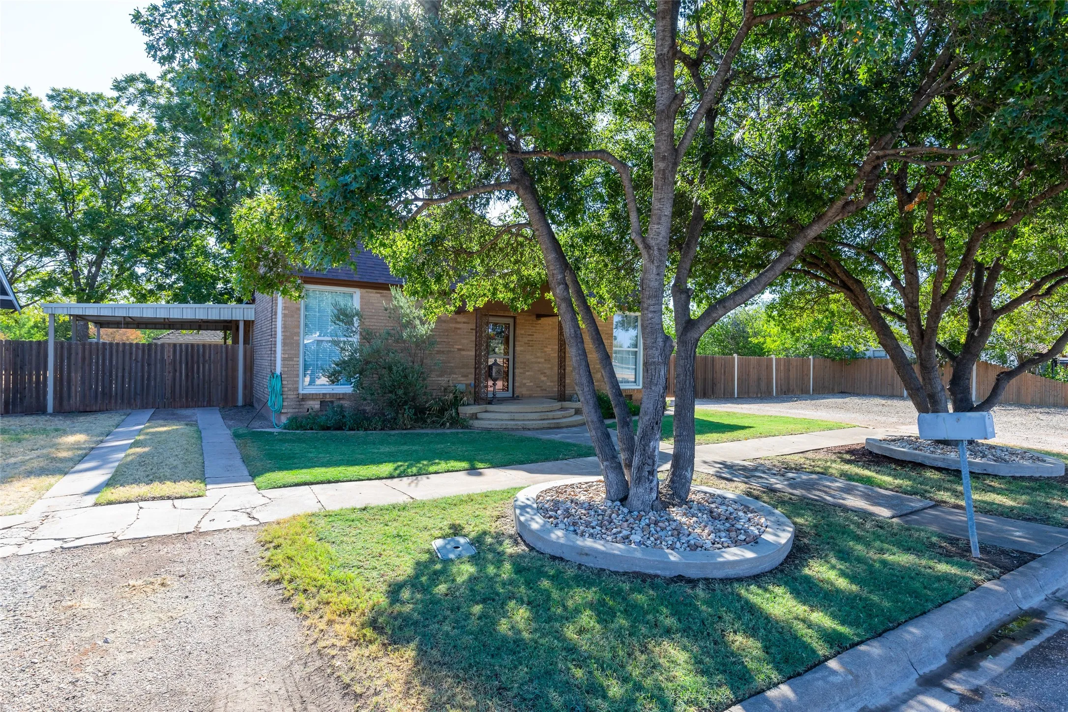 Brick exterior featuring a covered carport, mature trees, a manicured lawn, and a sidewalk leading to the entrance