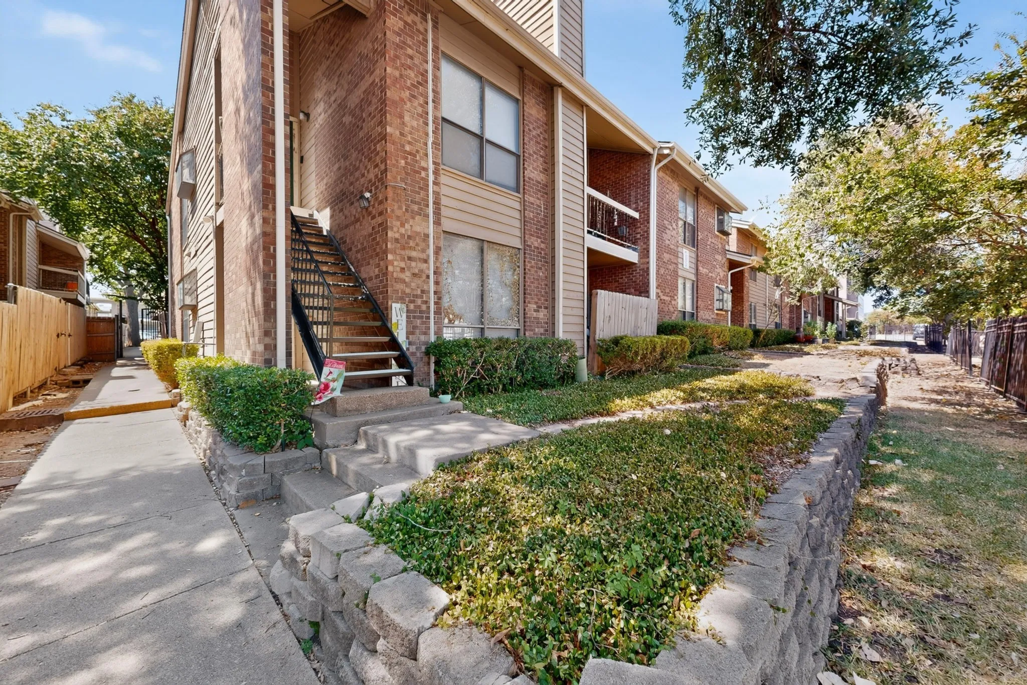 Brick exterior with vertical siding accents, concrete walkways, mature trees, and a metal staircase with railing