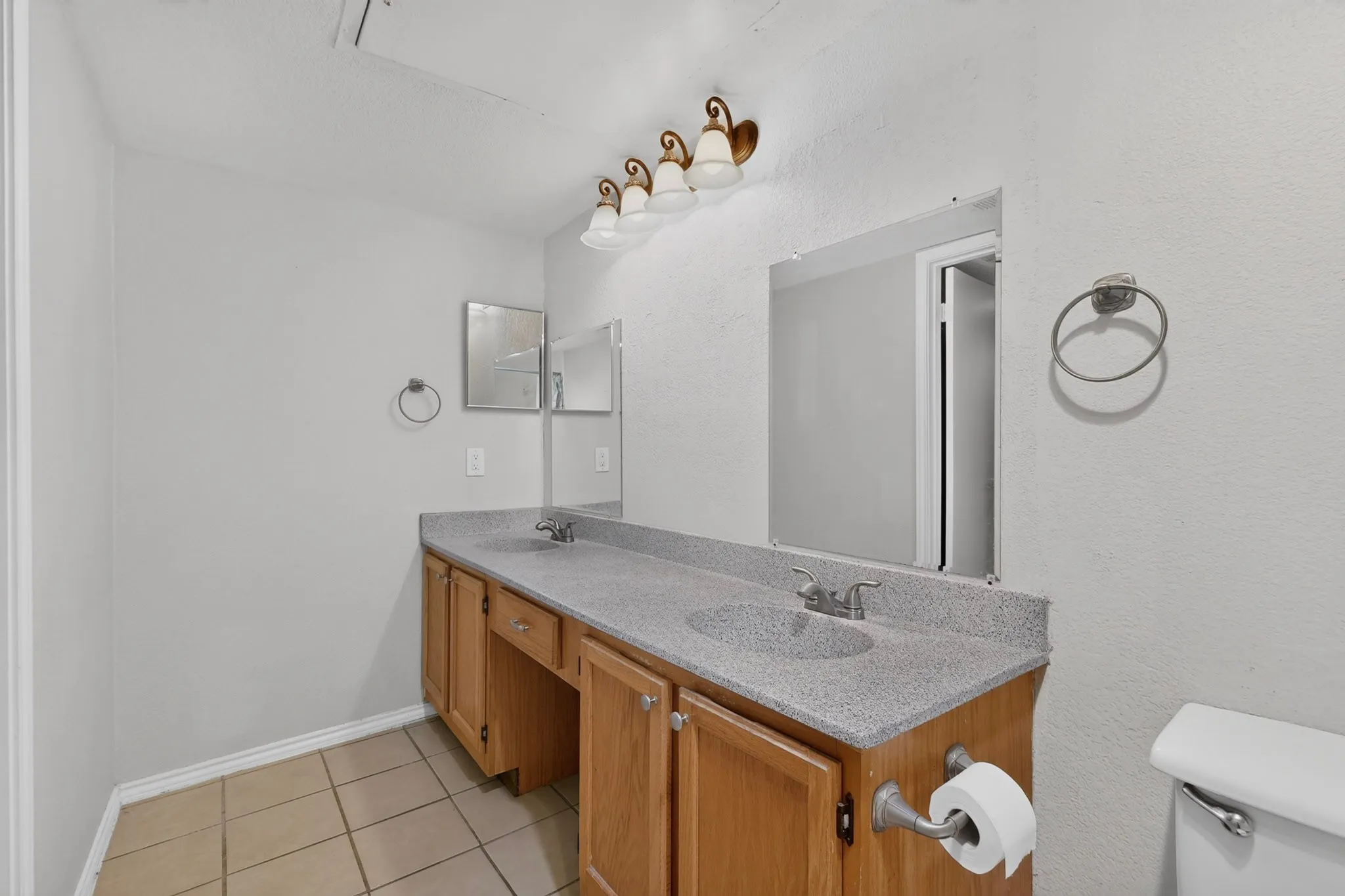 Dual sink vanity with wood-finish cabinetry, speckled countertop, and tiled flooring