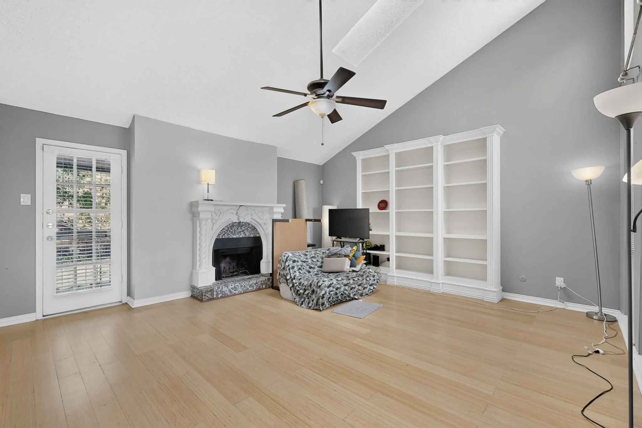 Vaulted ceiling, wood-finish flooring, ceiling fan, ornate fireplace with a marble-finish surround, and built-in shelving