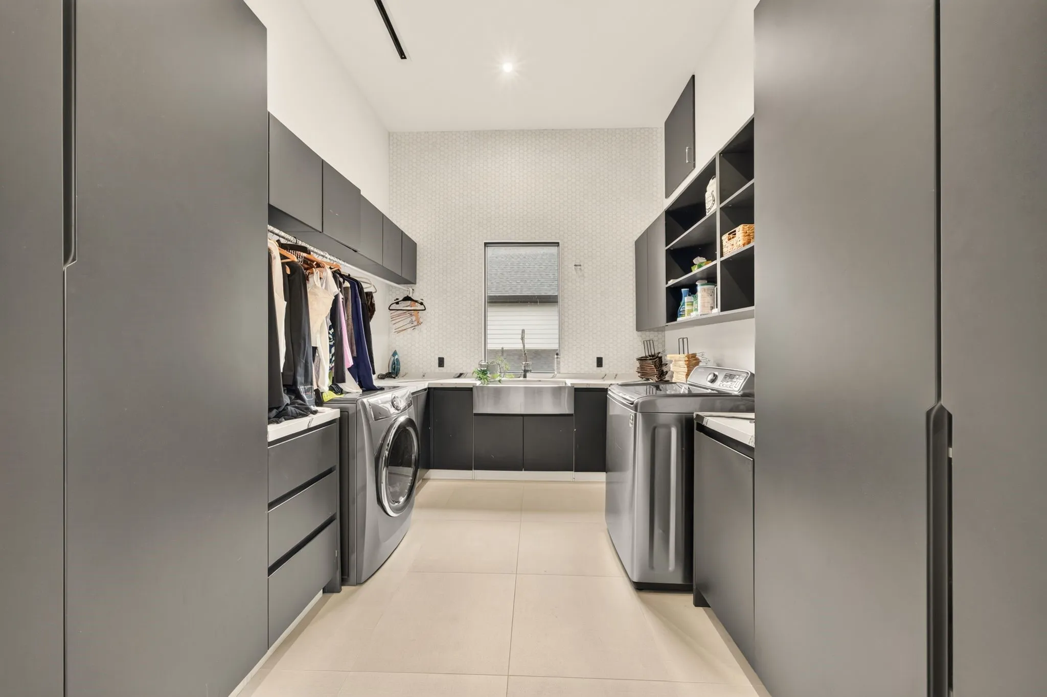 Specialized laundry room featuring a large stainless steel sink, extensive dark cabinetry, light hexagonal wall tiling, and a window with horizontal blinds