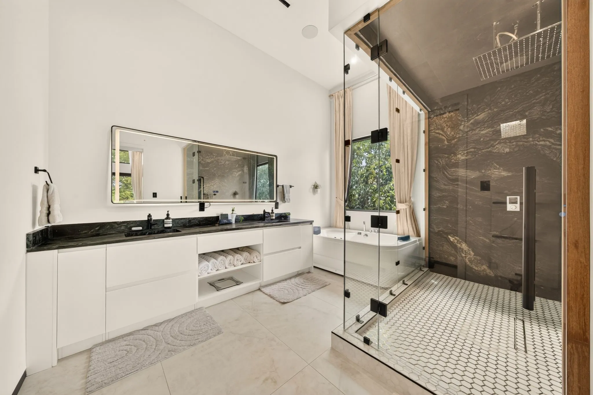 Dual vanity with dark countertop and white cabinetry, frameless glass shower enclosure with dark stone-finish walls, built-in soaking tub, and hexagon tile shower floor