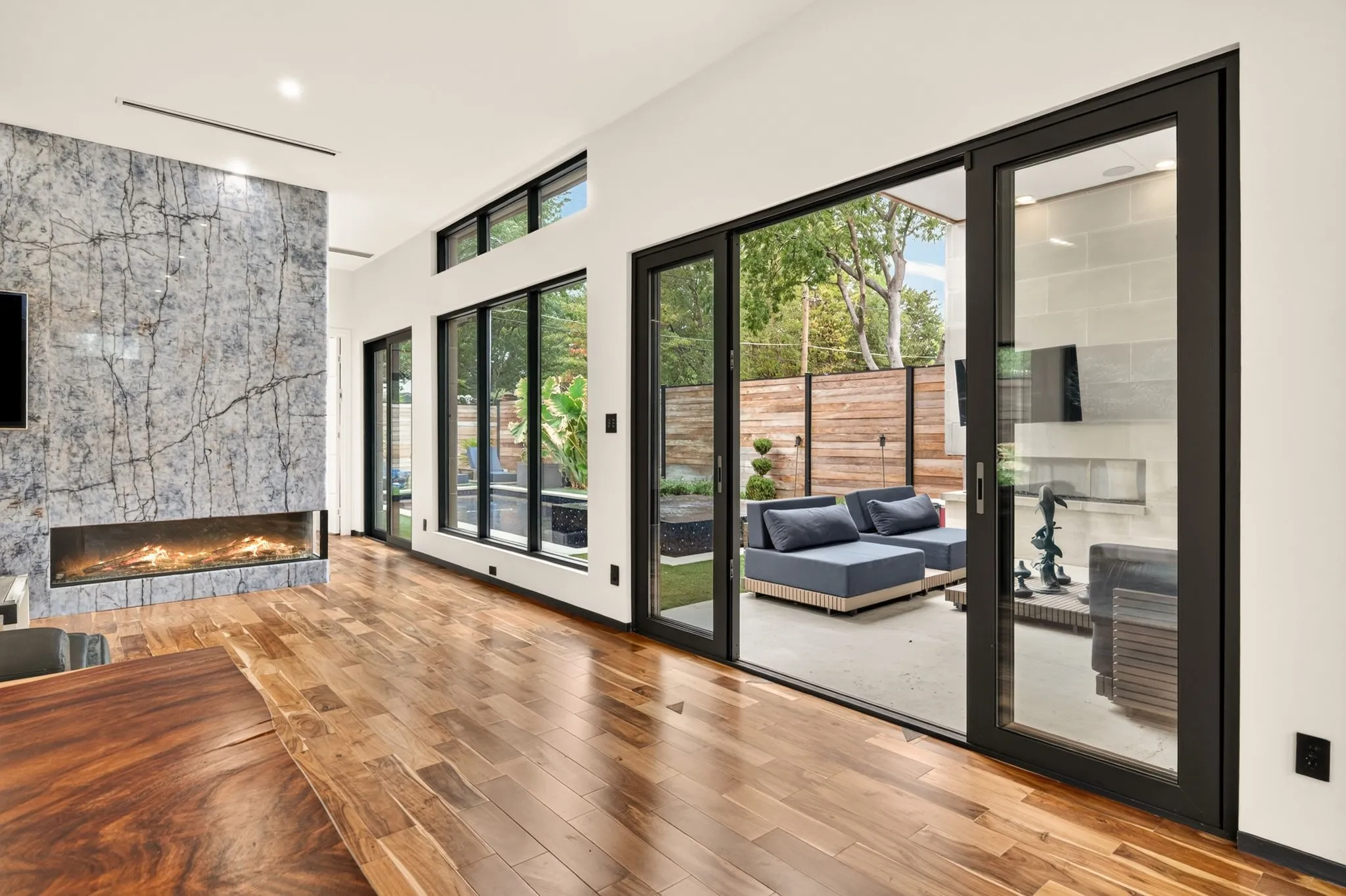 Hardwood flooring, full-height stone fireplace with linear firebox, large glass sliding doors, transom windows, and a paved outdoor patio