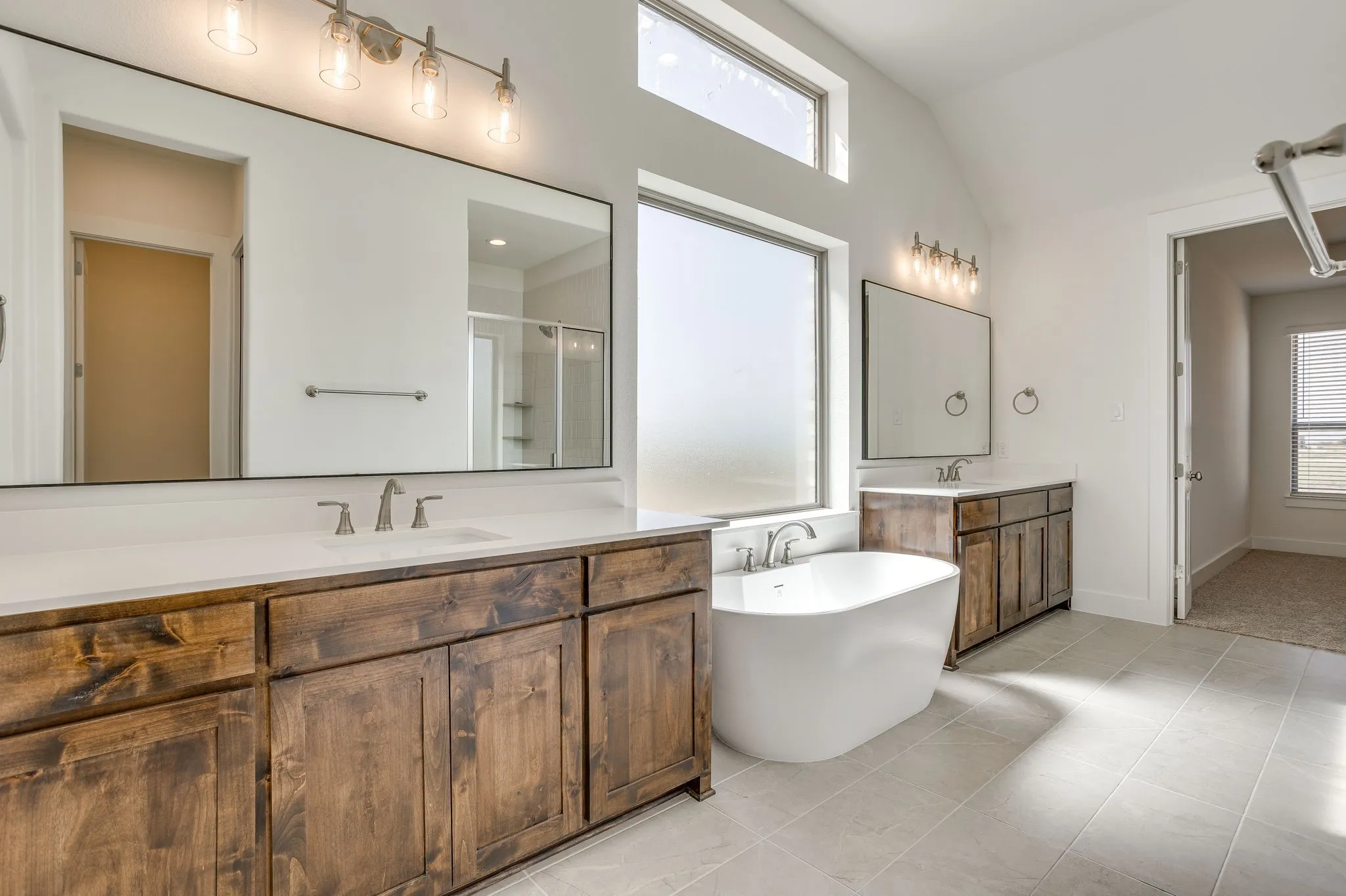 Soaking tub with a wide window, dual wood-finish vanities with undermount sinks, and light-toned tile flooring