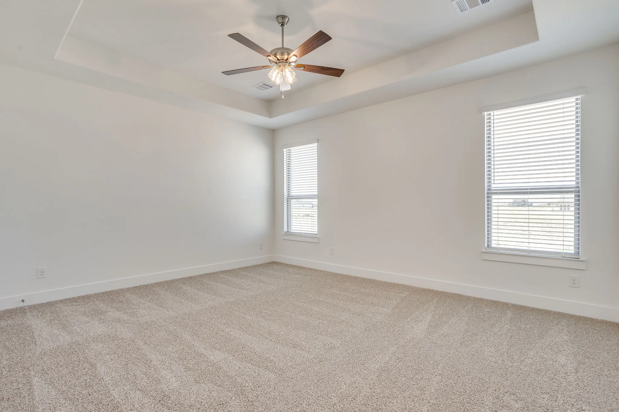 Tray ceiling with a ceiling fan, two windows with blinds, and neutral carpeting