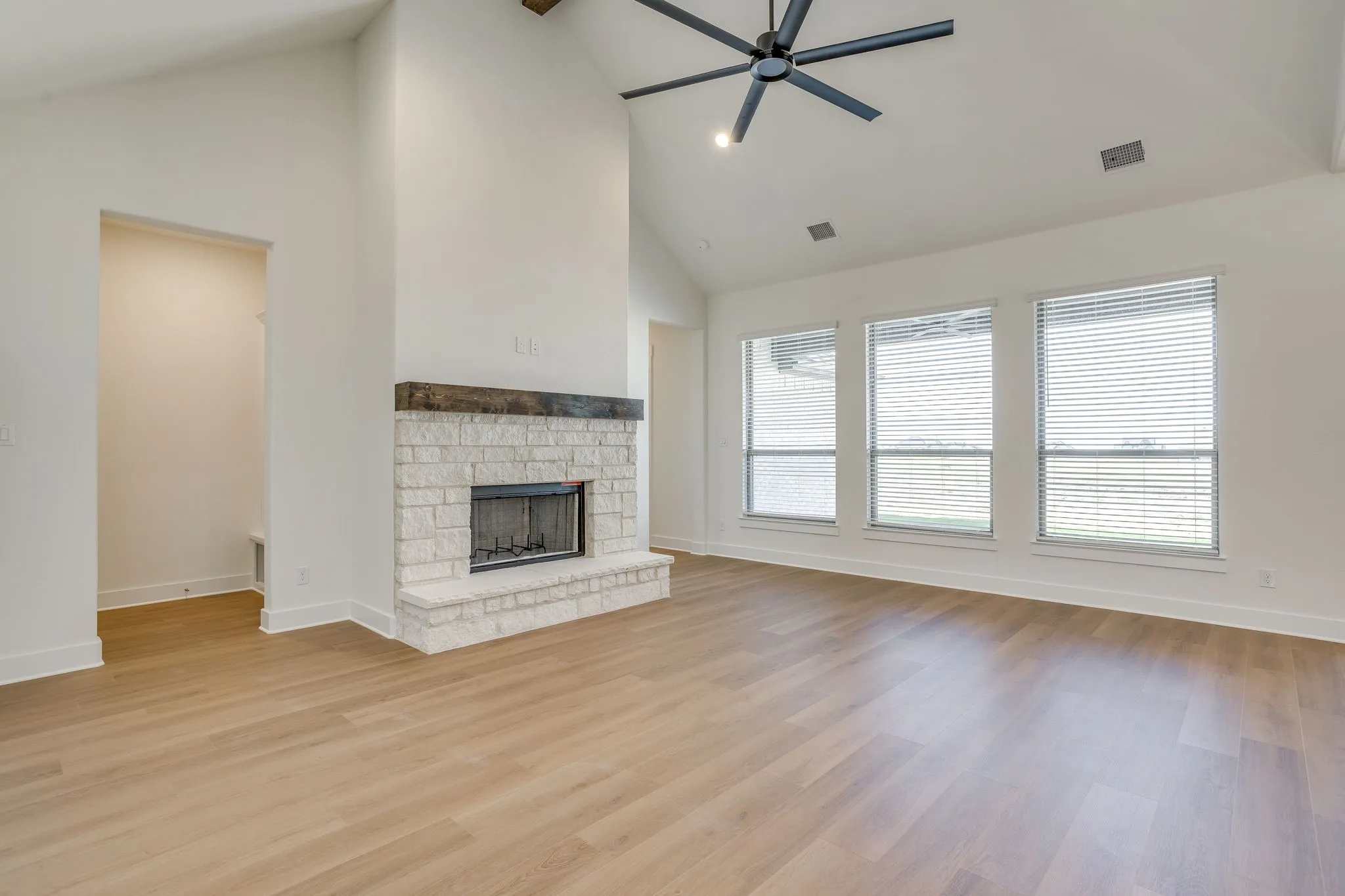Vaulted ceiling, stone-clad fireplace with wood mantel, wood-finish flooring, three large windows, and a ceiling fan