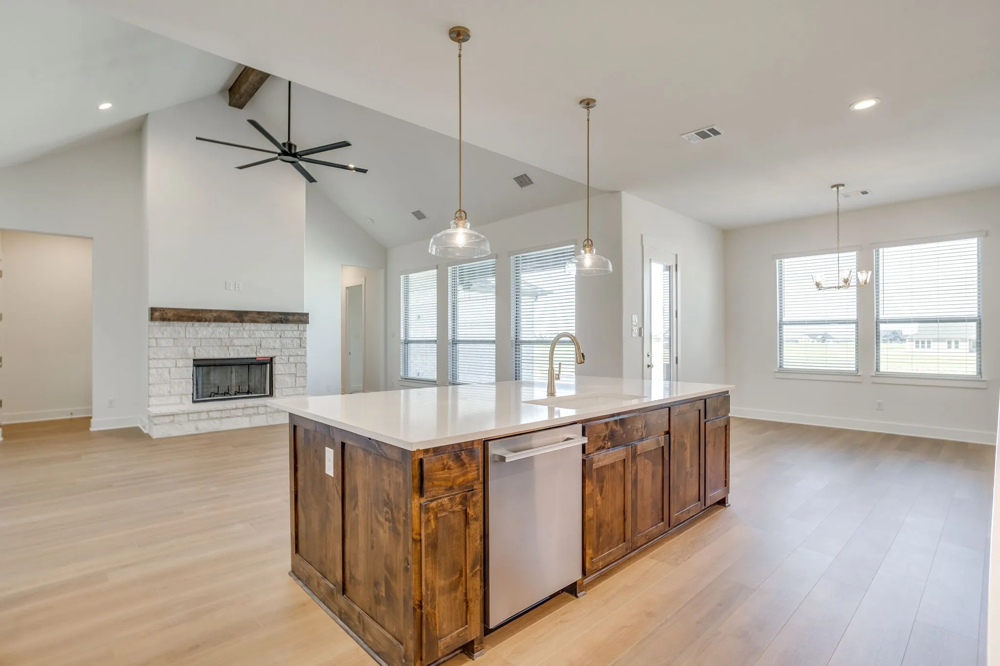Open-concept living space featuring wood-finish flooring, a kitchen island with stone countertop, stainless steel dishwasher, and a stone fireplace with a wood-beam mantel