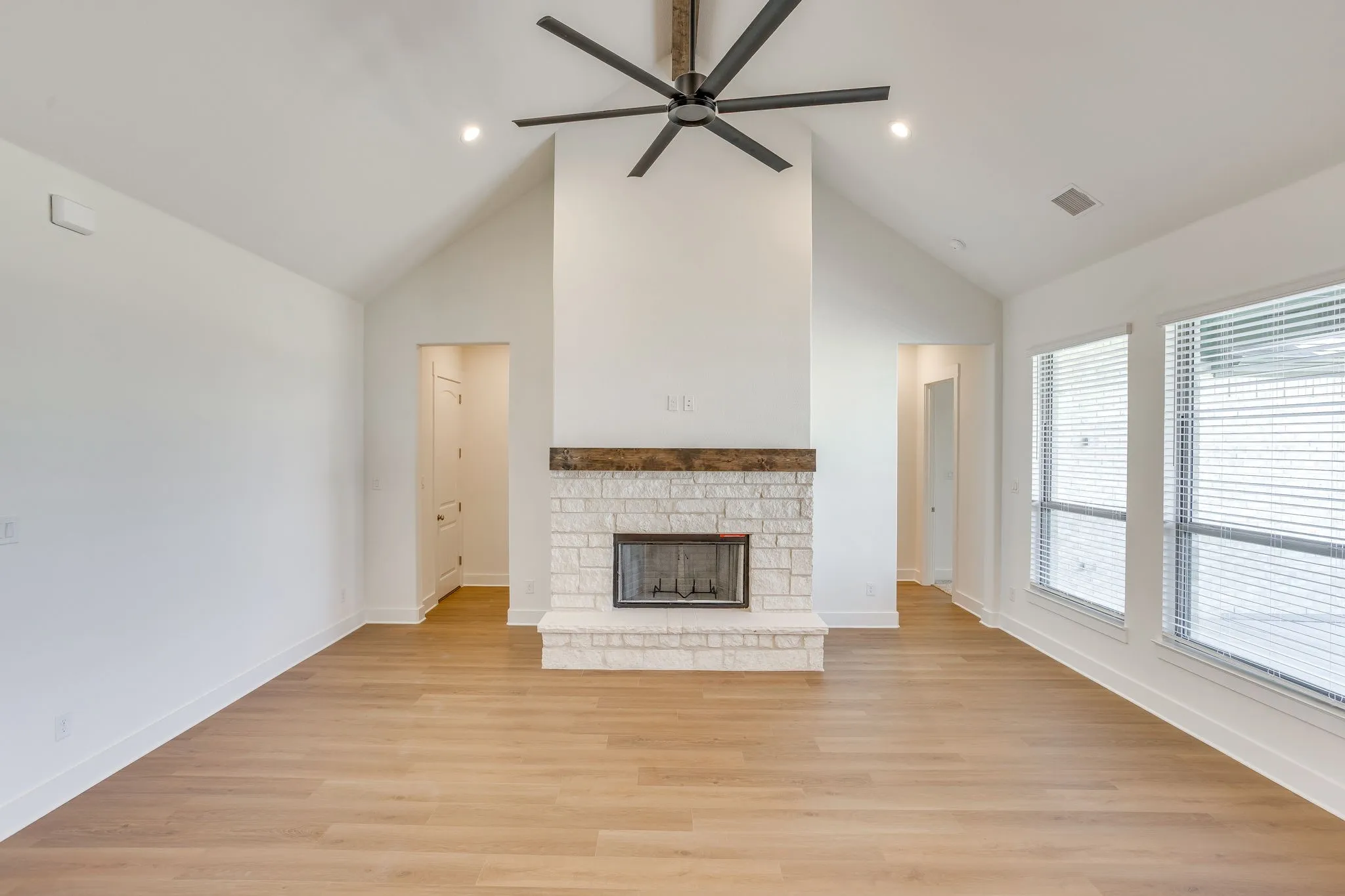 Vaulted ceiling with a ceiling fan, stone-clad fireplace with a wood mantle, wood-finish flooring, and multiple windows