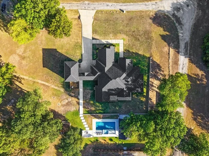 Spacious property featuring a large residence, concrete driveway, lush trees, and a private swimming pool with surrounding hardscaping