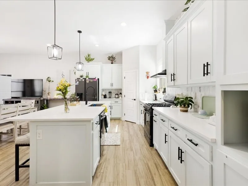 White cabinetry with matte black hardware, light-toned wood-finish flooring, white countertop surfaces, kitchen island with integrated sink, and pendant lighting