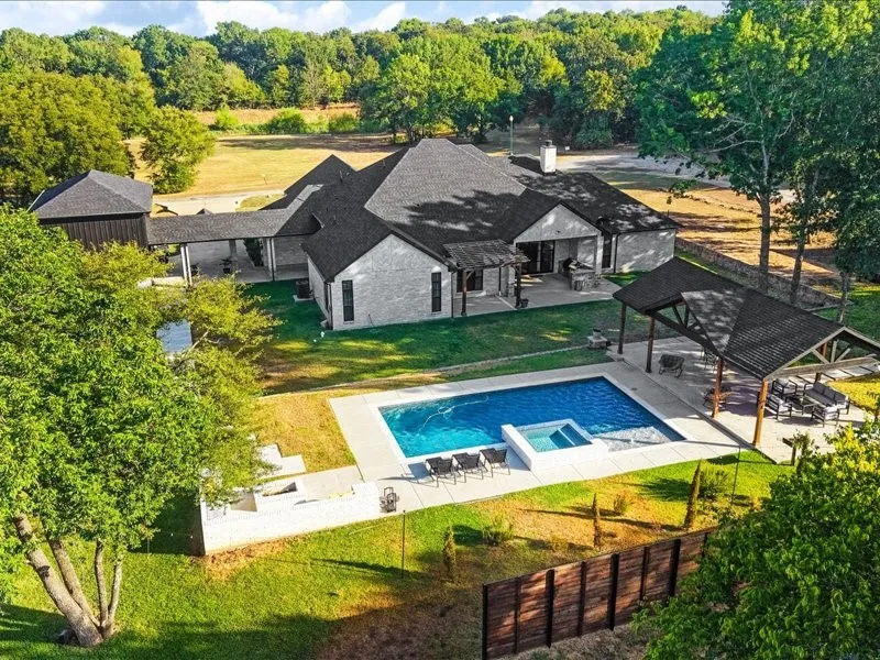 Exterior featuring light-colored brick with dark trim windows, dark shingle roof, swimming pool with integrated spa, covered outdoor living area, and a wood privacy fence