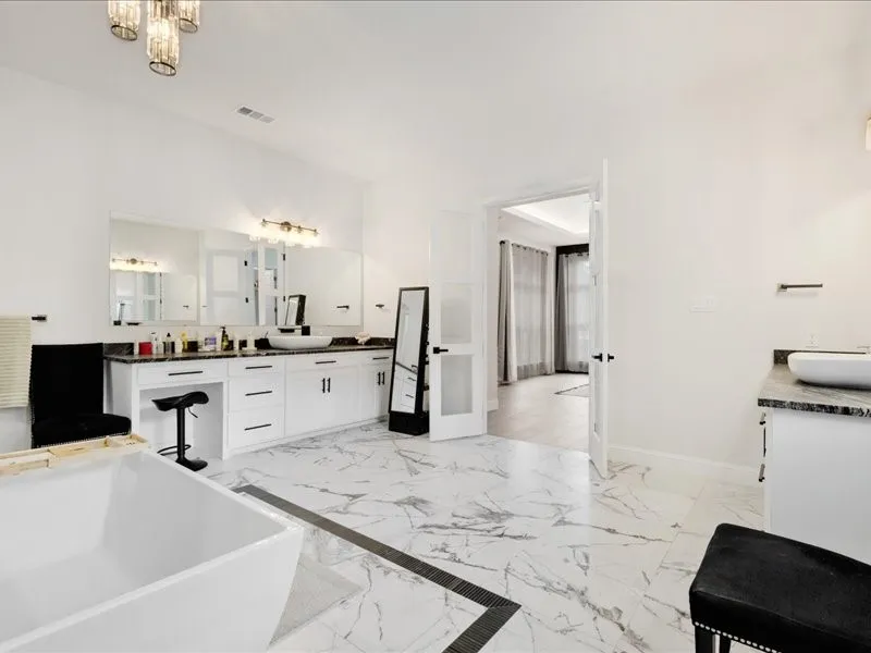 Freestanding soaking tub, white dual vanity with vessel sinks, marble-style tile flooring, and a crystal-fixture chandelier
