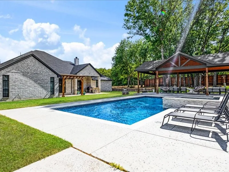 In-ground swimming pool with stone surround, expansive patio, detached covered pavilion with wood beams, and a wood pergola