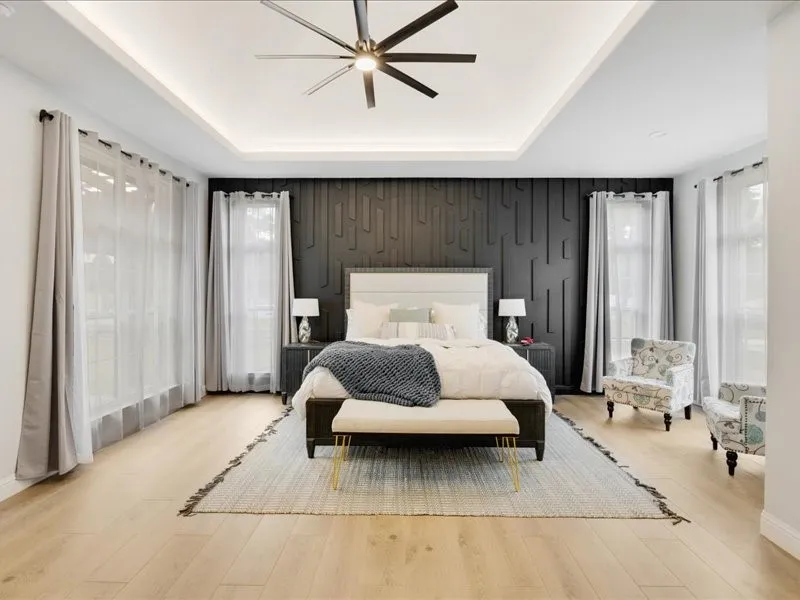 Tray ceiling with recessed lighting, modern ceiling fan, accent wall with geometric paneling, light wood-finish flooring, and multiple windows