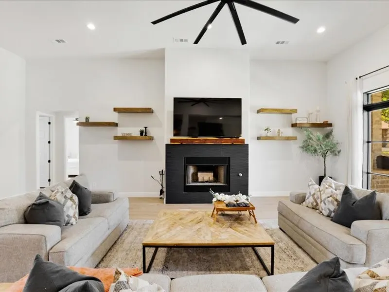 Vaulted ceiling with a contemporary ceiling fan, recessed lighting, black tiled fireplace with wood mantle, light wood-finish flooring, and built-in floating shelves