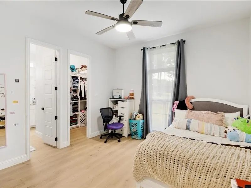 Wood-finish flooring, ceiling fan with integrated lighting, window with drapes, and a built-in closet organization system