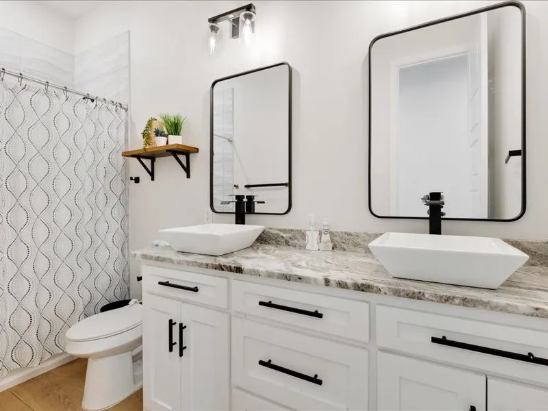 Dual vessel sinks with individual mirrors, white shaker-style cabinetry, a stone-finish countertop, and wood-finish flooring