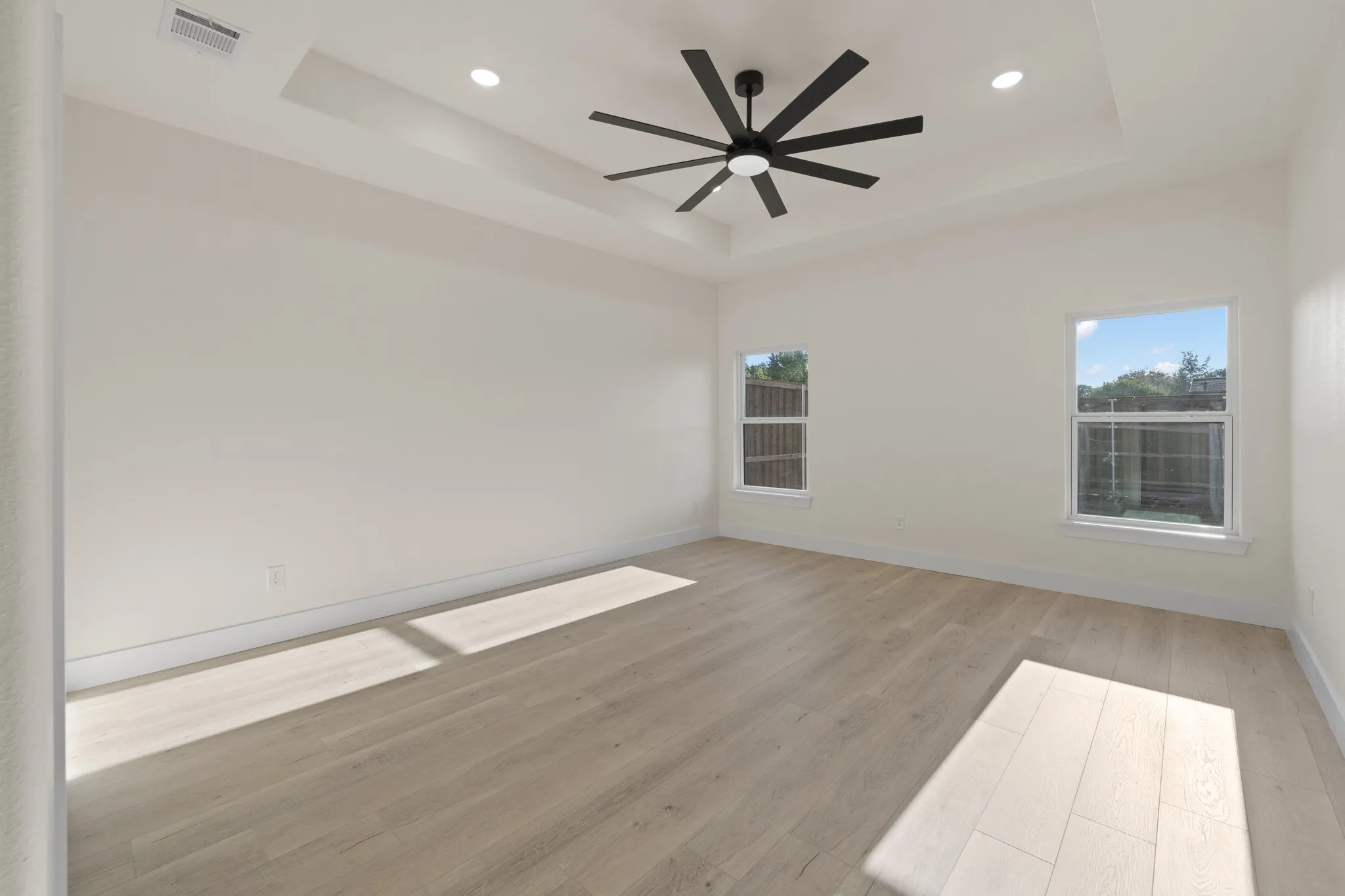 Spacious and inviting master bedroom featuring a beautiful tray ceiling with a modern oversized ceiling fan and recessed lighting. Large windows fill the room with natural light, while the light-toned flooring and neutral walls create a bright, contemporary feel.