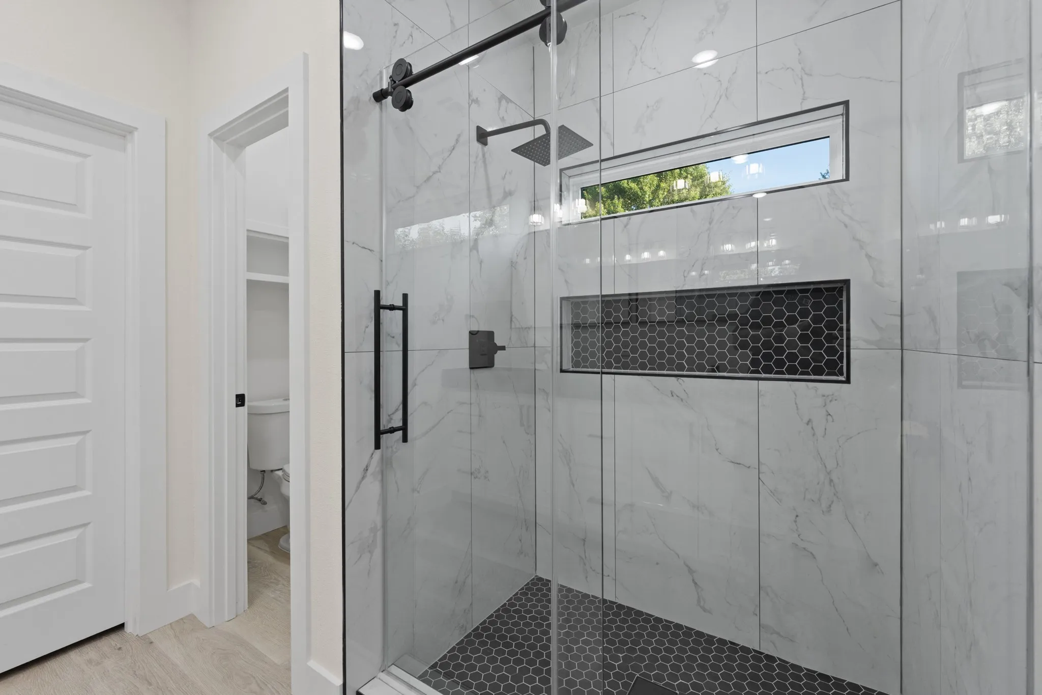 Glass-enclosed shower with black hardware, white tile walls, a horizontal window, and a recessed hexagonal tile niche