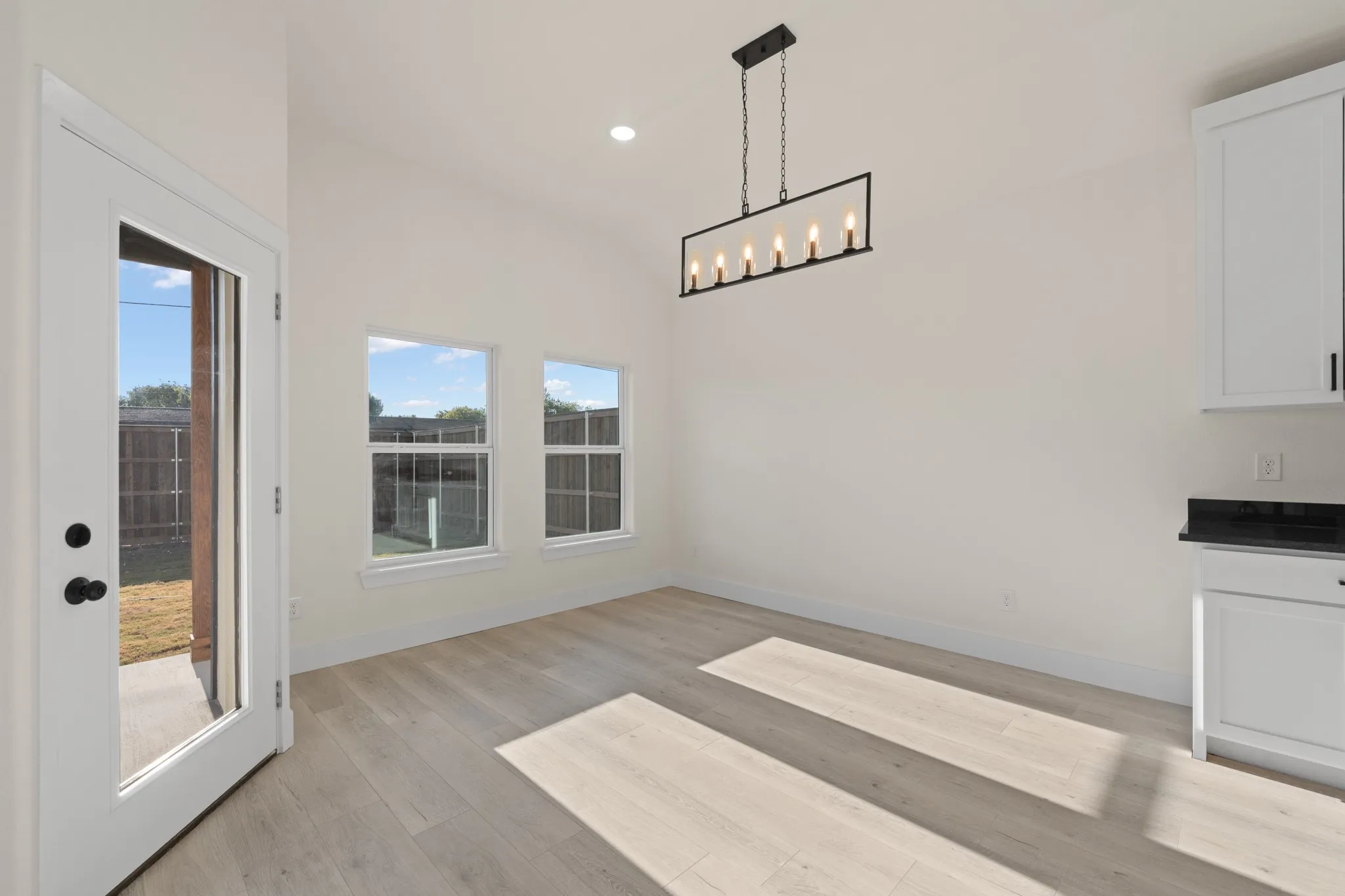 Vaulted ceiling with recessed lighting, wood-finish flooring, two large windows, white cabinetry with black hardware, and a black countertop