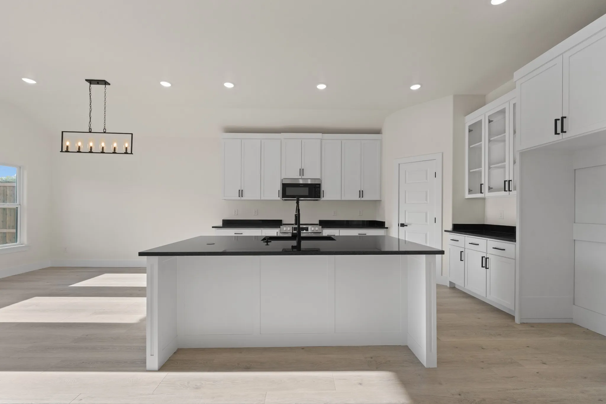 White cabinetry with matte black hardware, dark countertops, a kitchen island with an integrated sink, recessed lighting, and wood-finish flooring