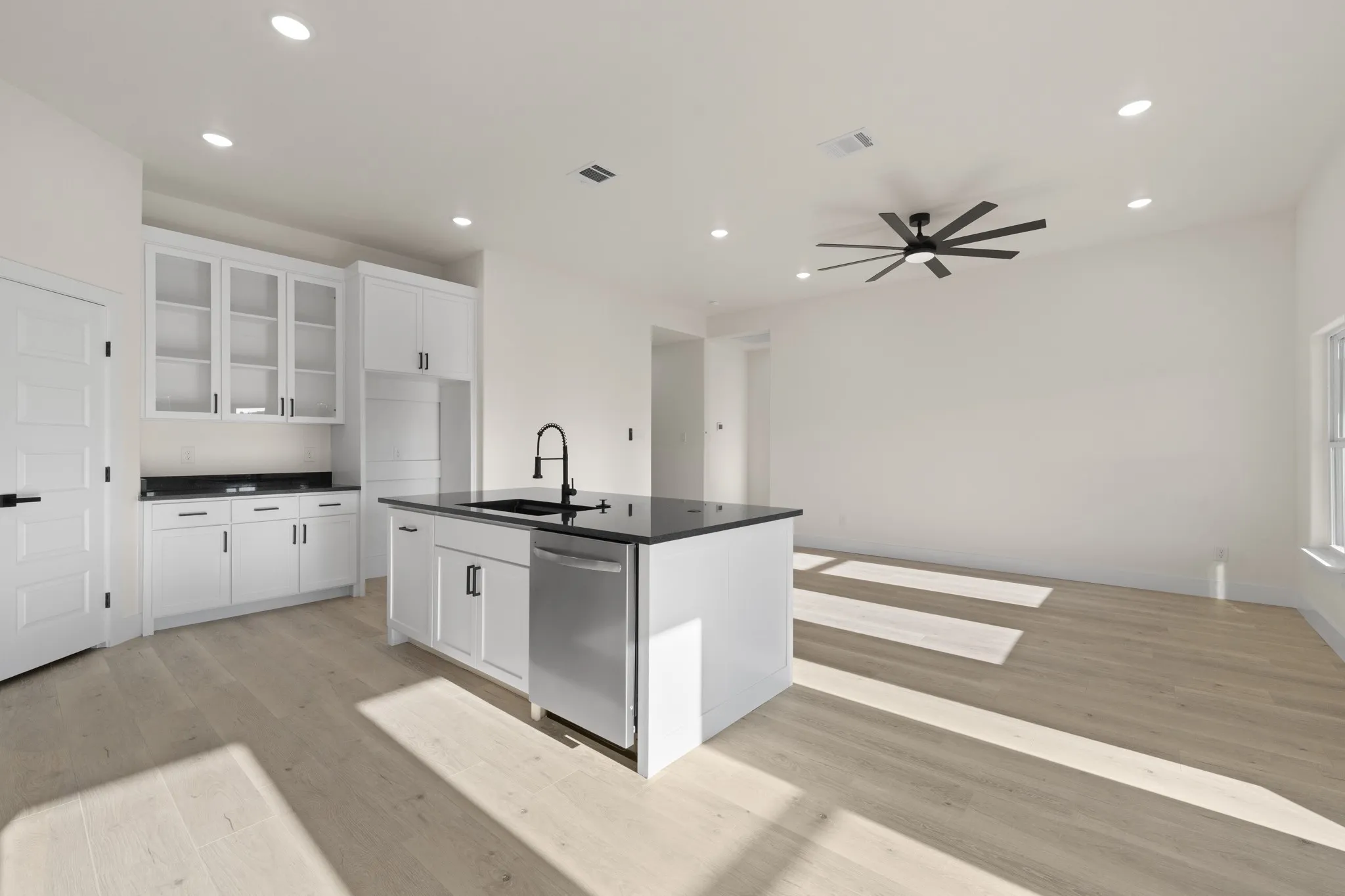 Modern kitchen featuring white cabinetry, black countertops, a central island with a built-in sink, stainless steel dishwasher, and wood-finish flooring