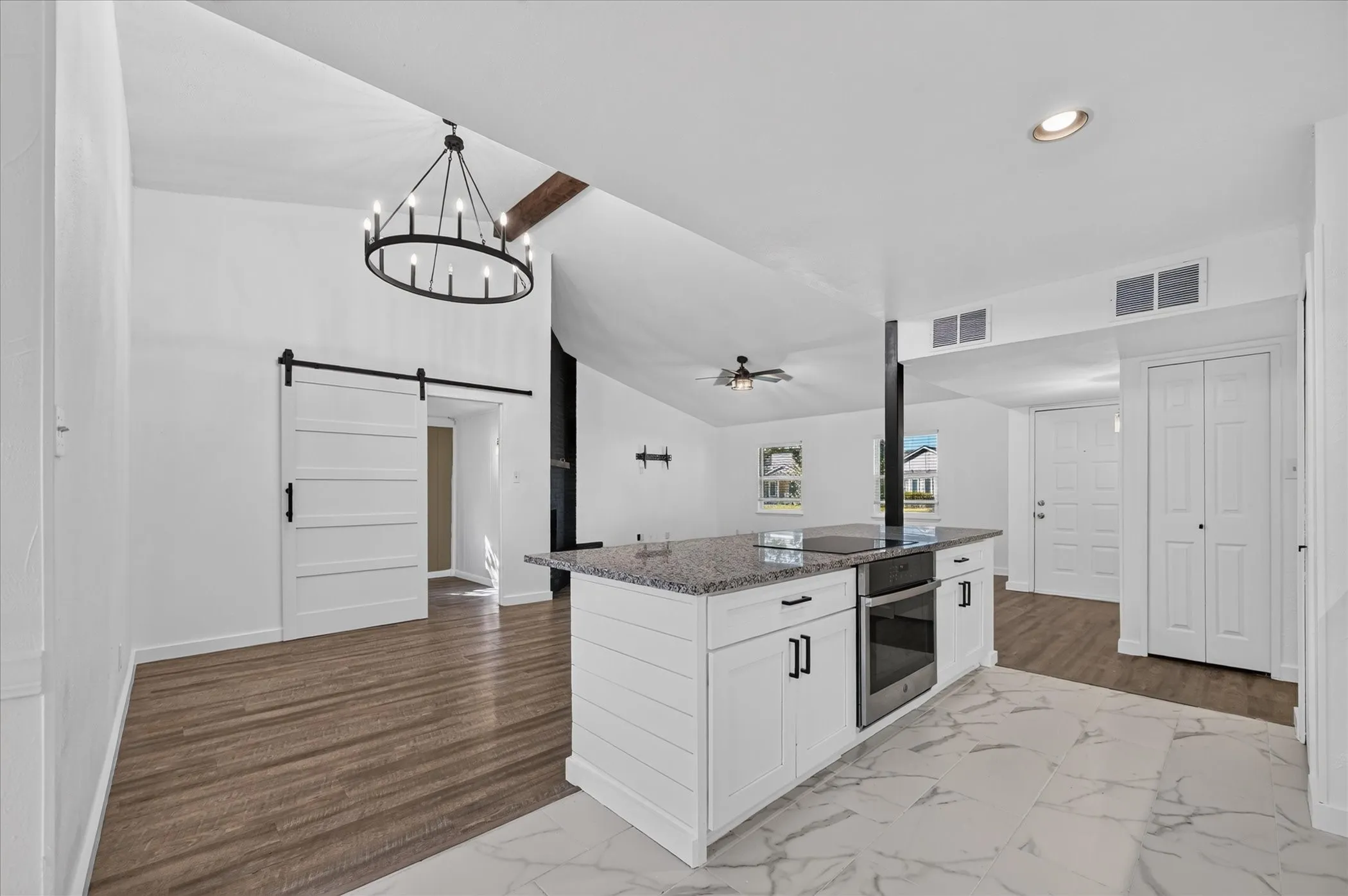 Vaulted ceiling with an exposed beam, kitchen island with granite countertop and built-in oven, white shaker cabinetry with black hardware, wood-finish flooring, and a sliding barn door