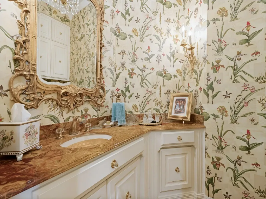 Ornate gold-framed mirror, botanical-patterned wallpaper, built-in cabinetry, polished stone countertop, and brass fixtures