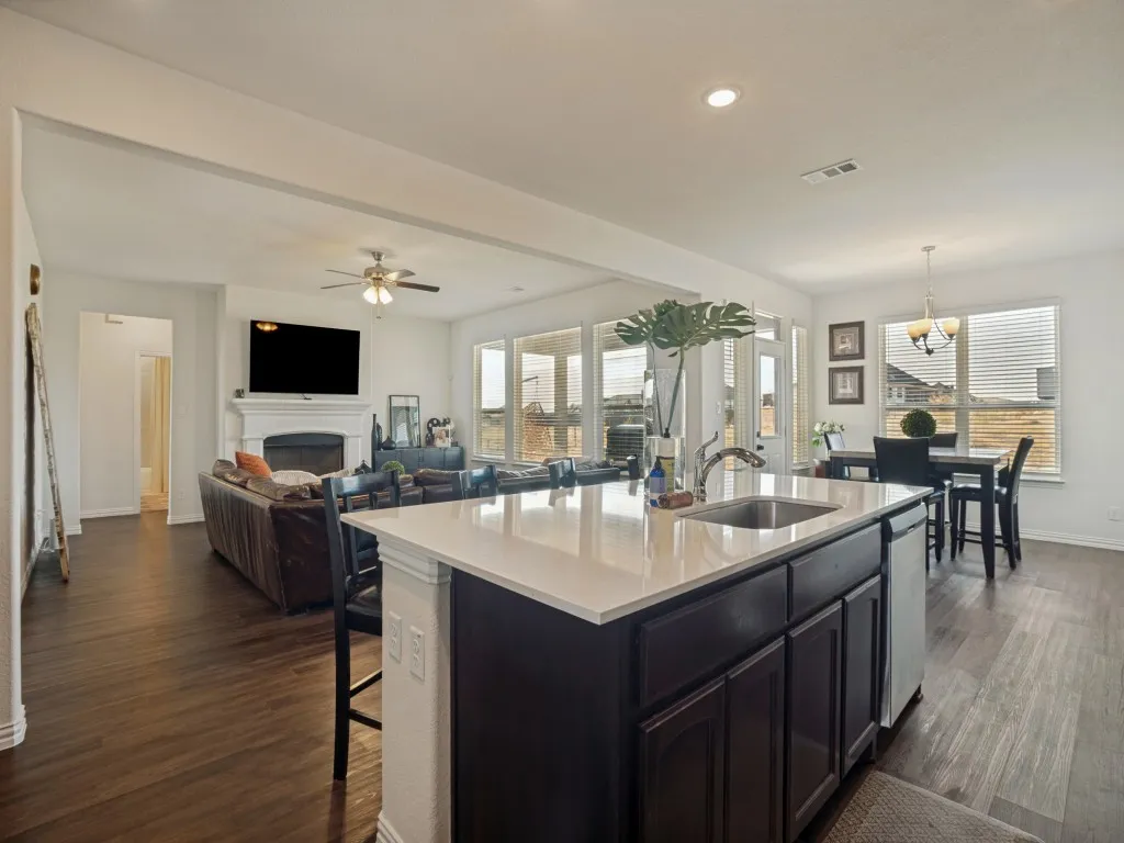 Open concept layout featuring wood-finish flooring, a kitchen island with sink, a fireplace with a white mantel, and ample natural light from multiple windows