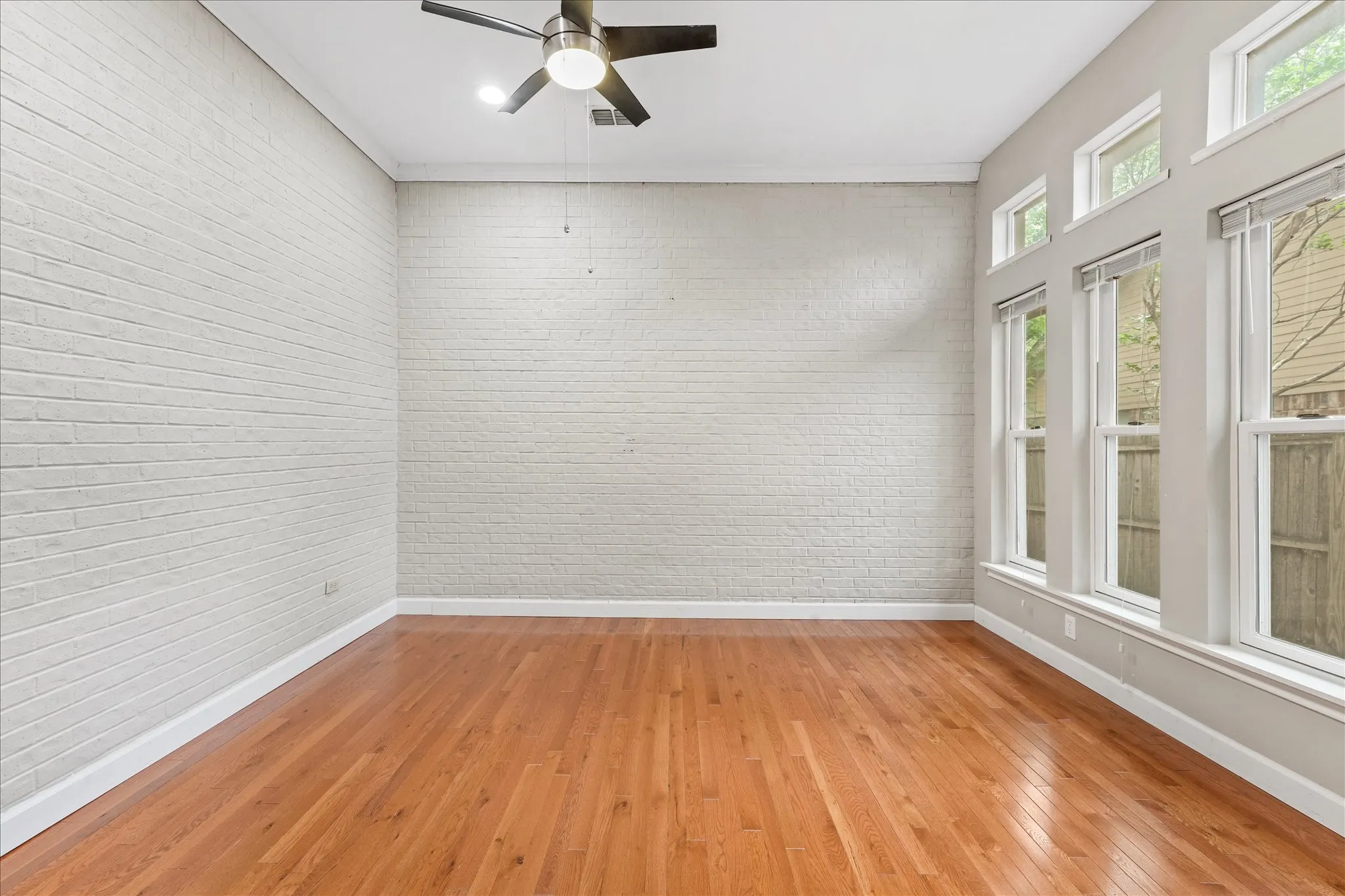 Two-story living room with soaring ceilings, painted brick fireplace, hardwood flooring, and oversized windows bringing in natural light and tree-lined views.