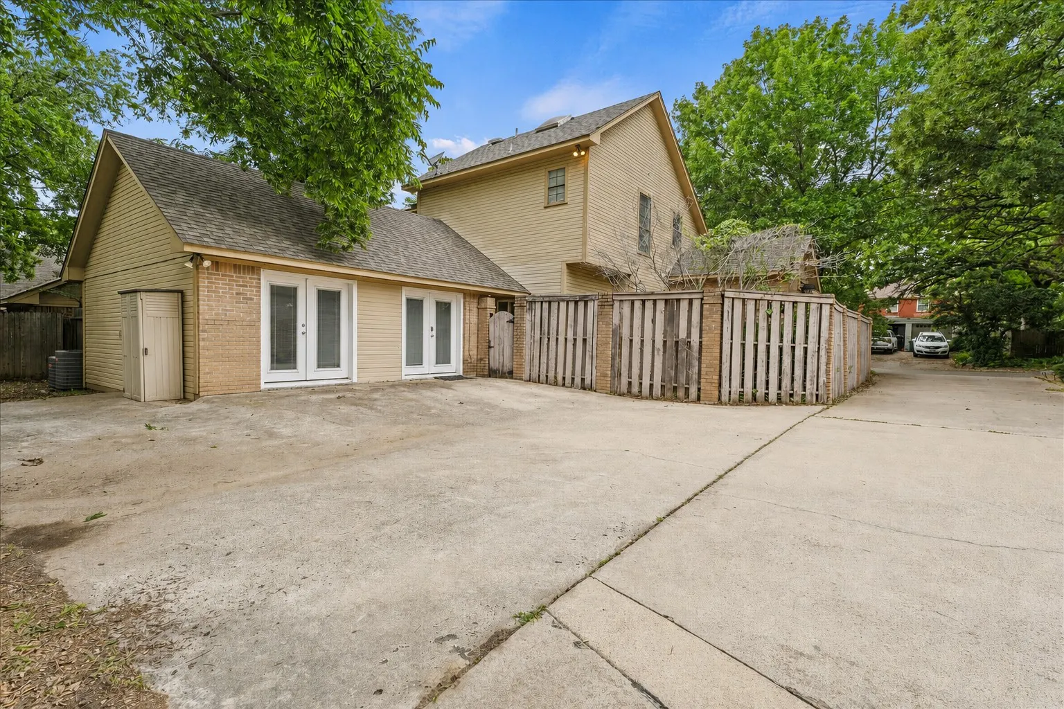 Rear exterior with private gated courtyard entry, multiple French doors, mature shade trees, and oversized driveway with additional parking space.