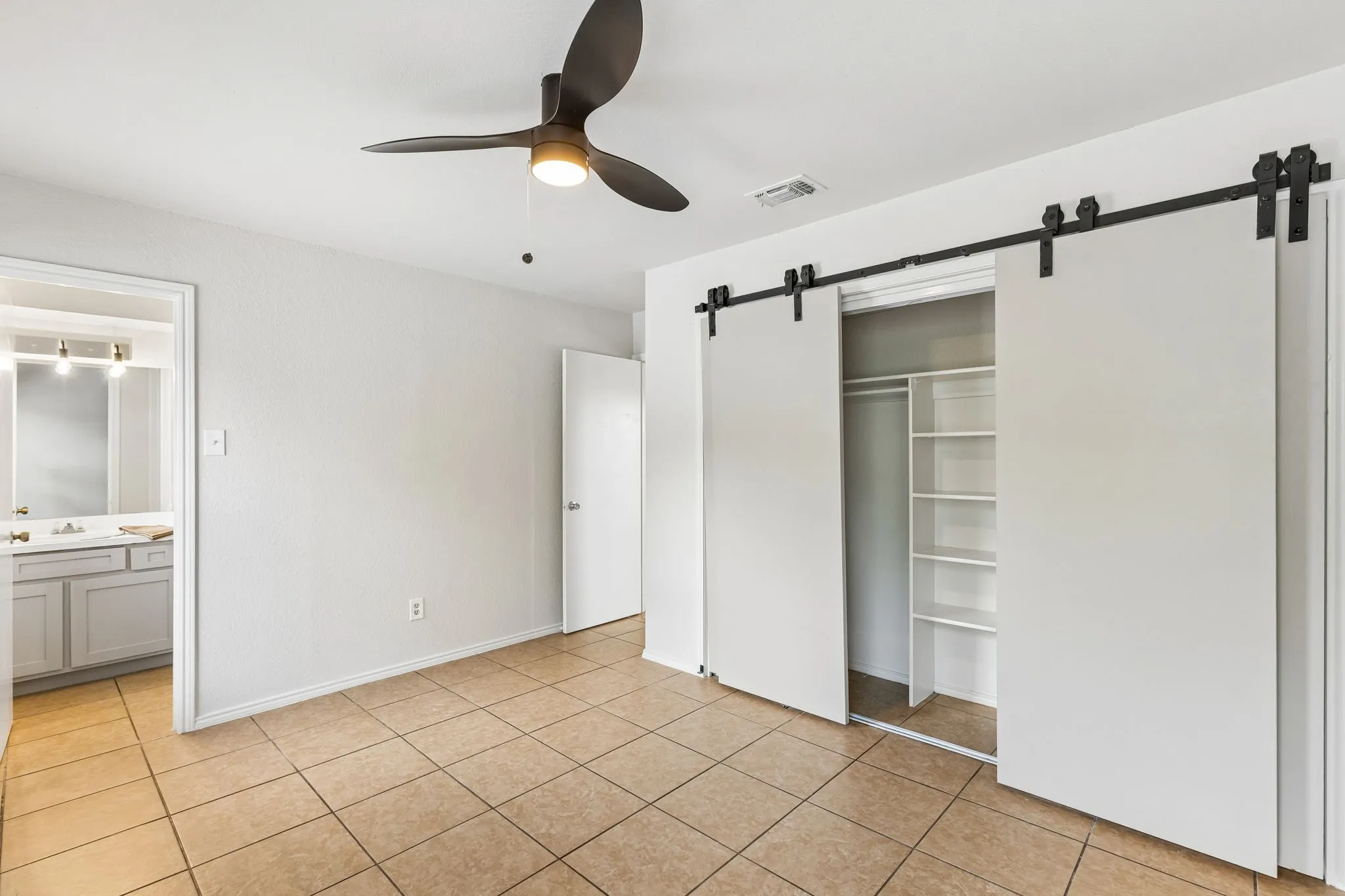 Tile flooring throughout, modern ceiling fan with integrated lighting, sliding barn doors for closet access, built-in closet shelving, and a vanity with a countertop sink in the attached bathroom