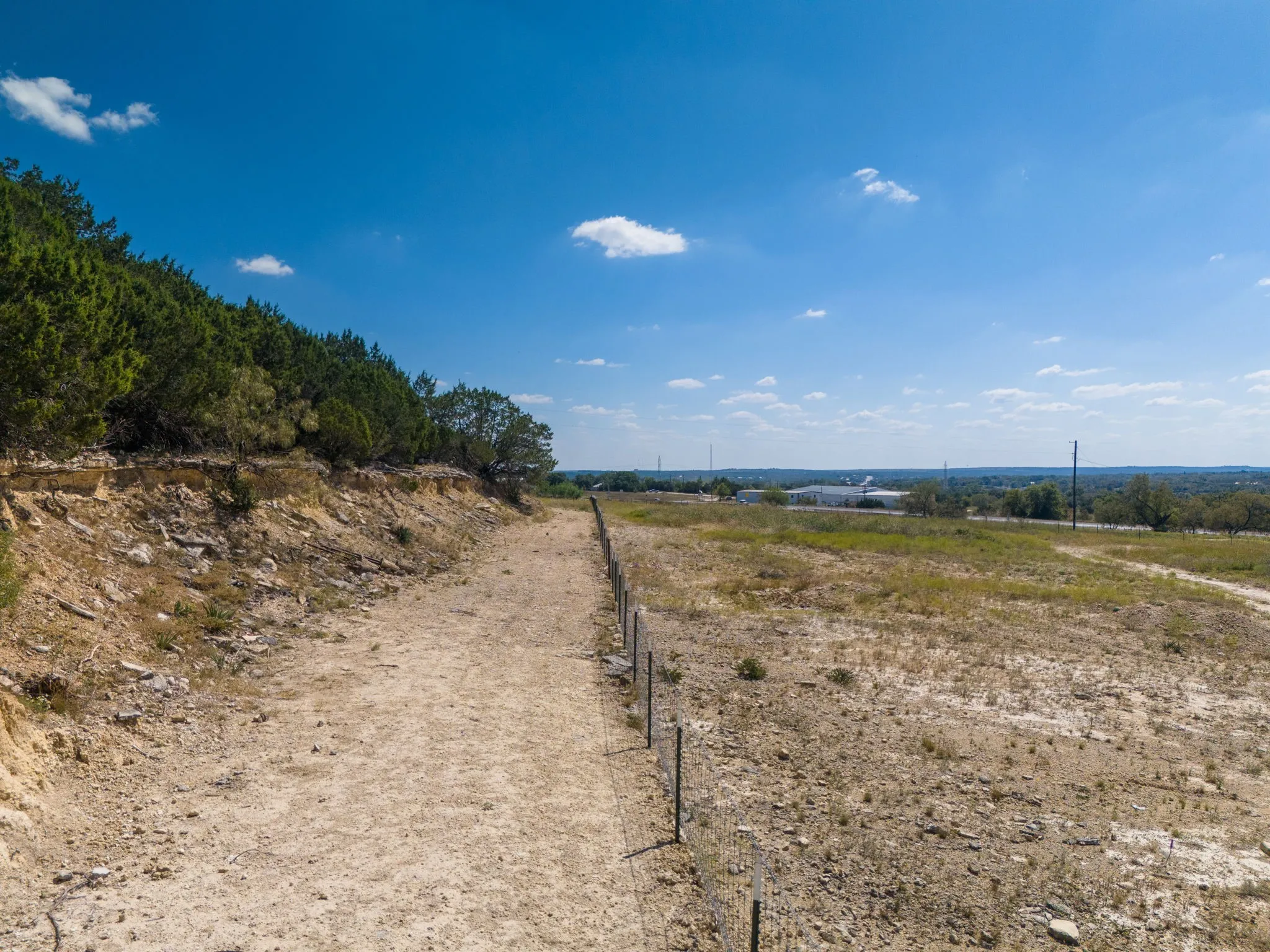 Expansive land parcel featuring natural terrain, a partial wire fence, and distant structures under a clear sky