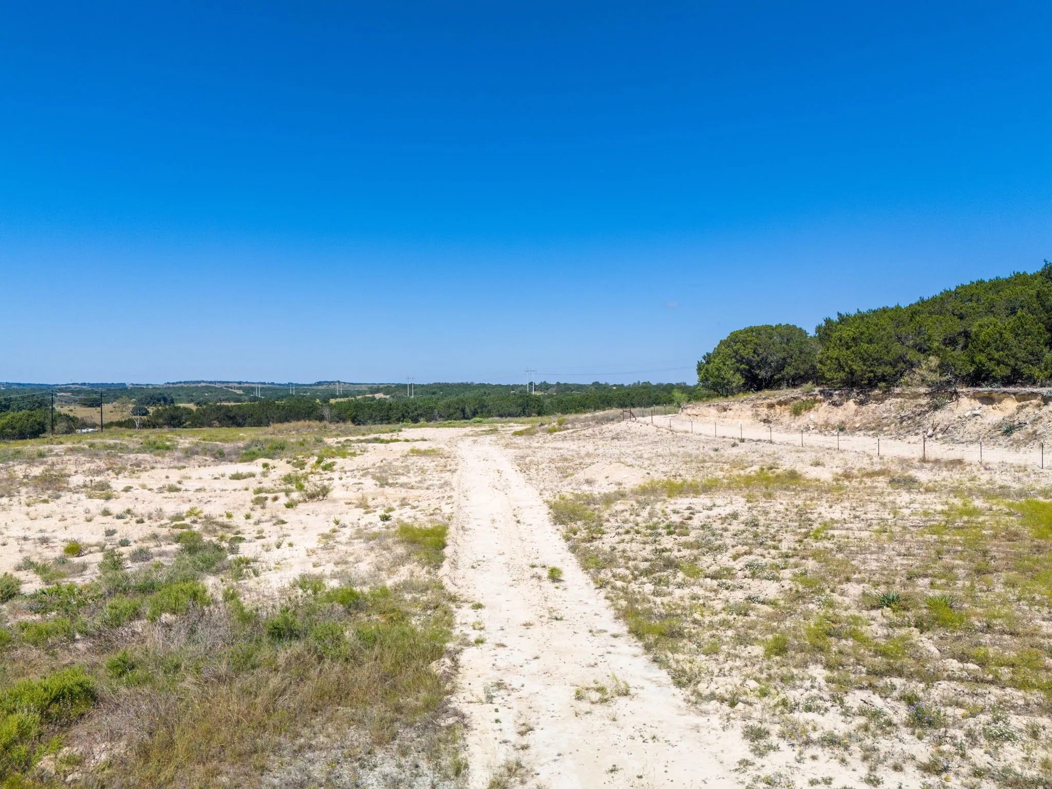 Expansive undeveloped land with a dirt access path, mature trees bordering the property, and distant natural terrain