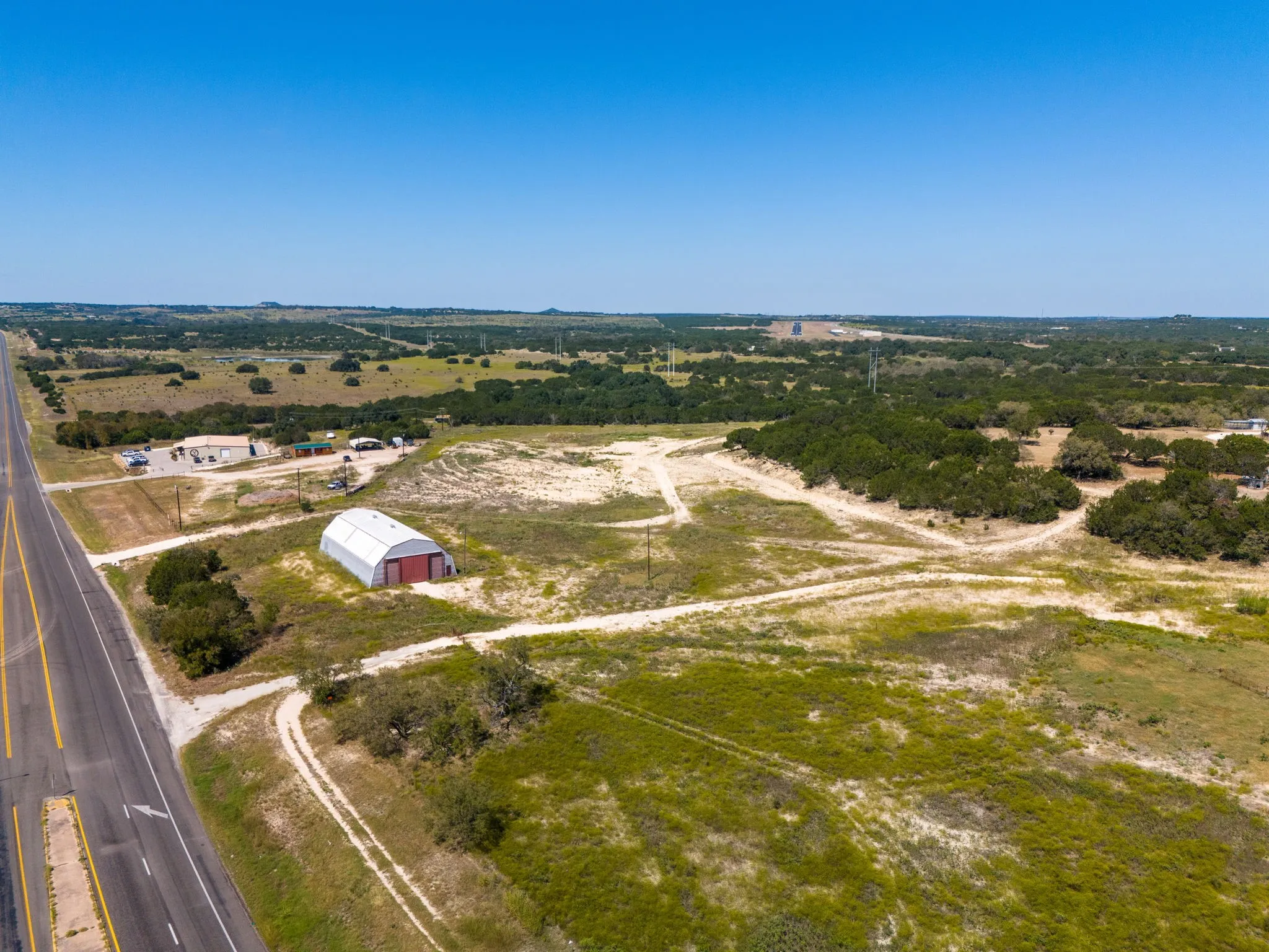 Expansive undeveloped land with a partial paved road border, featuring a large metal utility building with red doors, and a natural landscape with scattered trees