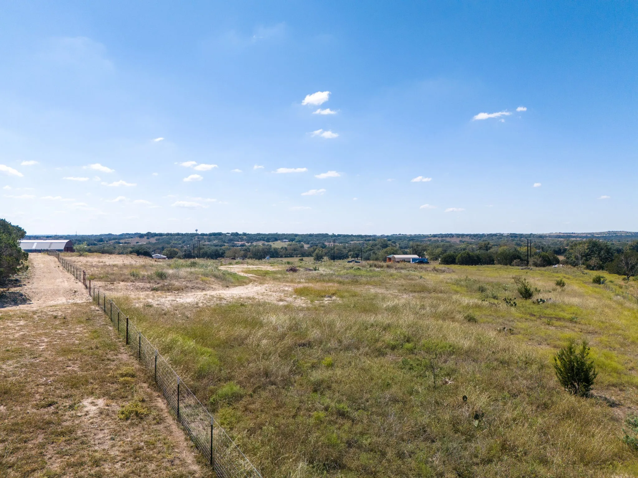 Expansive open land featuring natural grasses and mature trees, with a distant structural building and a perimeter fence