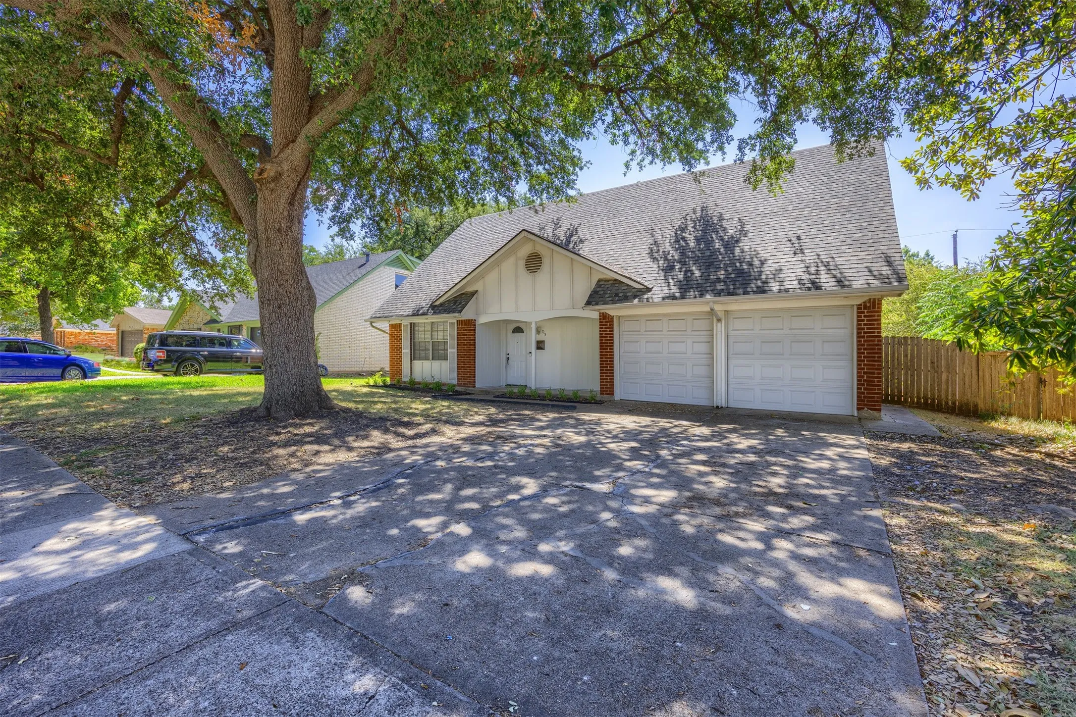 Gable roofline with asphalt shingles, two-car garage, brick facade accents, mature tree, and concrete driveway