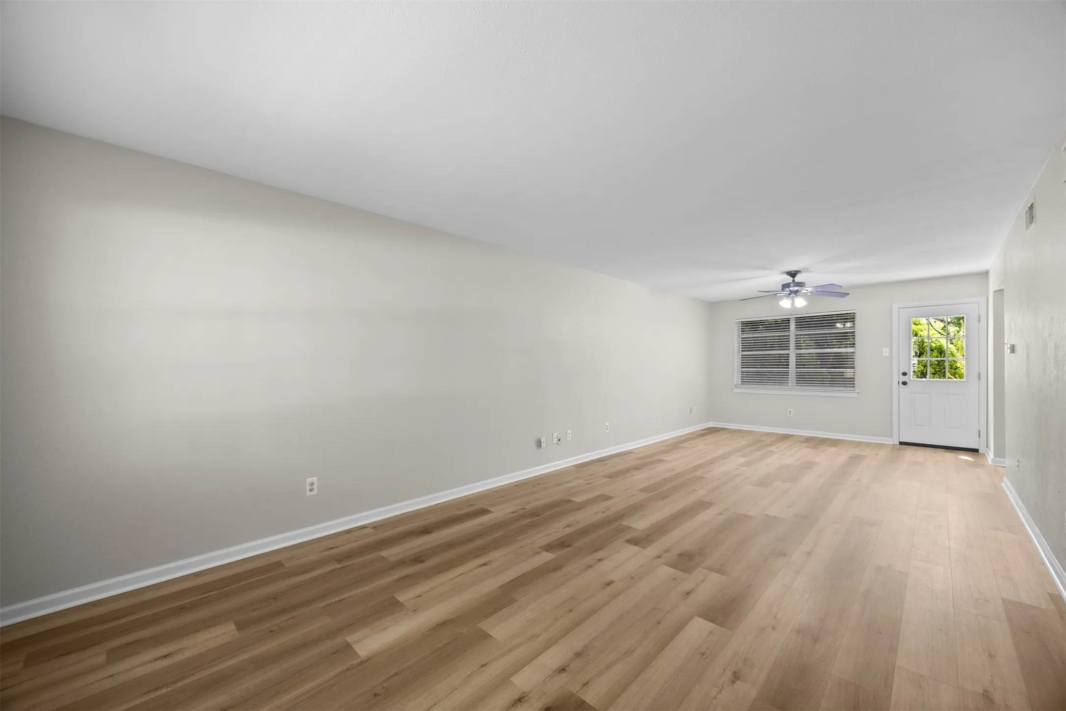 Spacious room featuring wood-finish flooring, a ceiling fan with integrated lighting, multiple windows, and a door with a glass insert