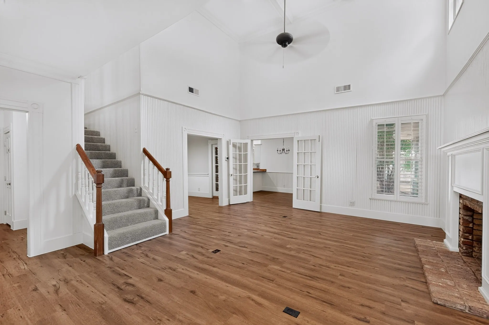 High vaulted ceiling with a ceiling fan, wood-finish flooring, white shiplap wall paneling, brick fireplace with a white mantel, and a carpeted staircase with wood handrails