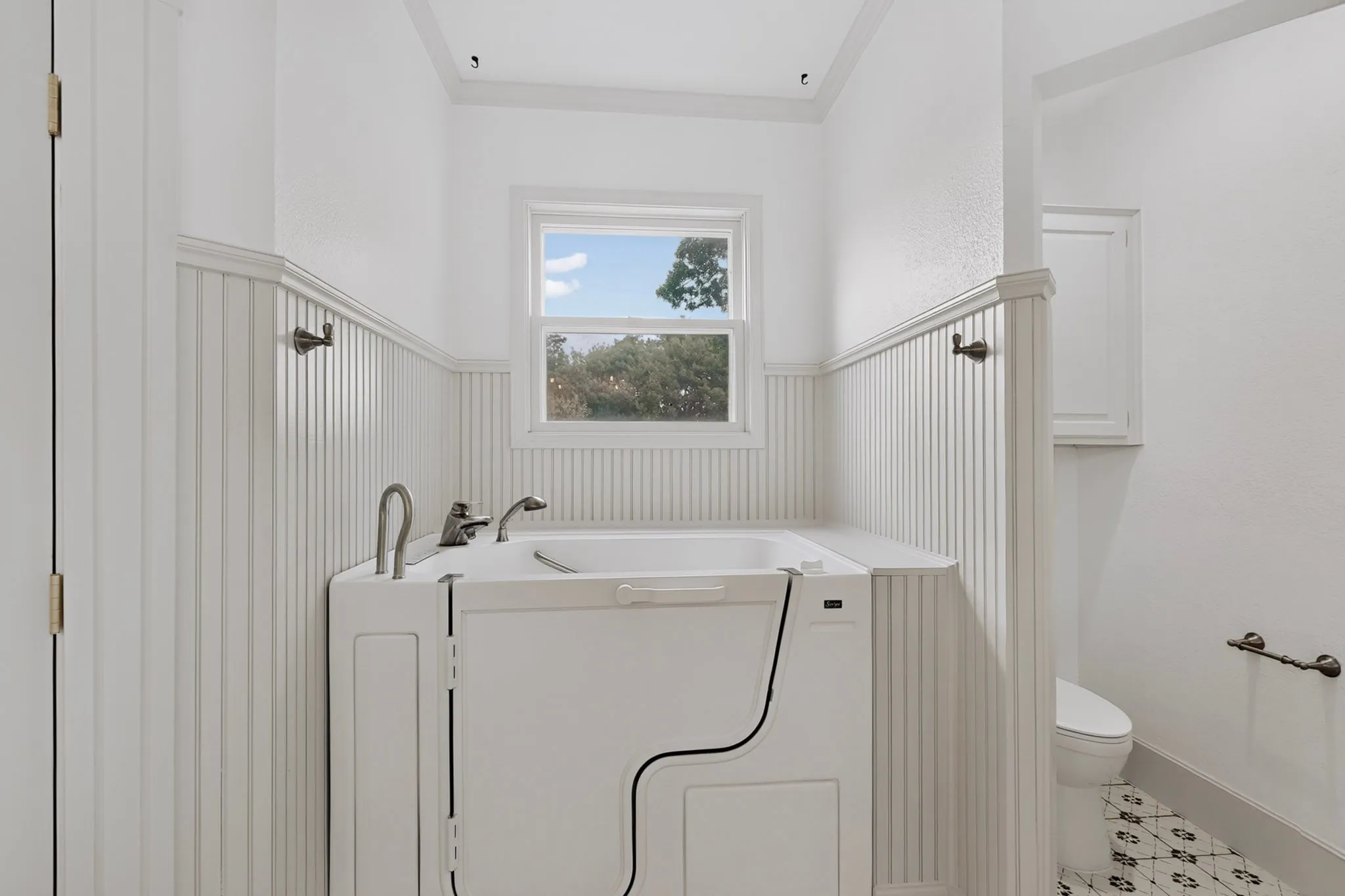 Walk-in bathtub with chrome fixtures, wainscoting, window with exterior scenery, and patterned tile flooring