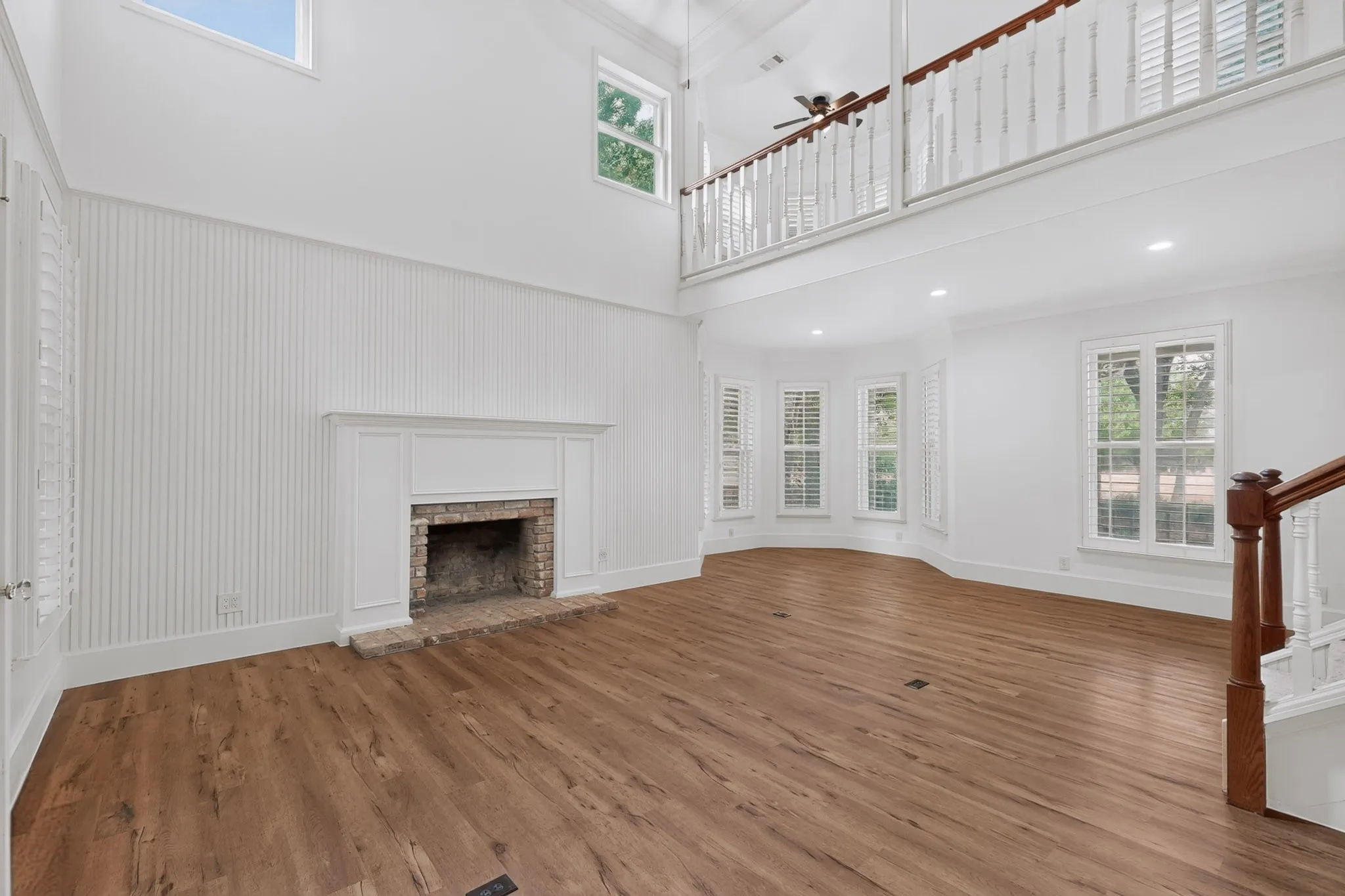 Two-story living area with wood-finish flooring, a brick fireplace with a white mantel, and a bay window featuring plantation shutters