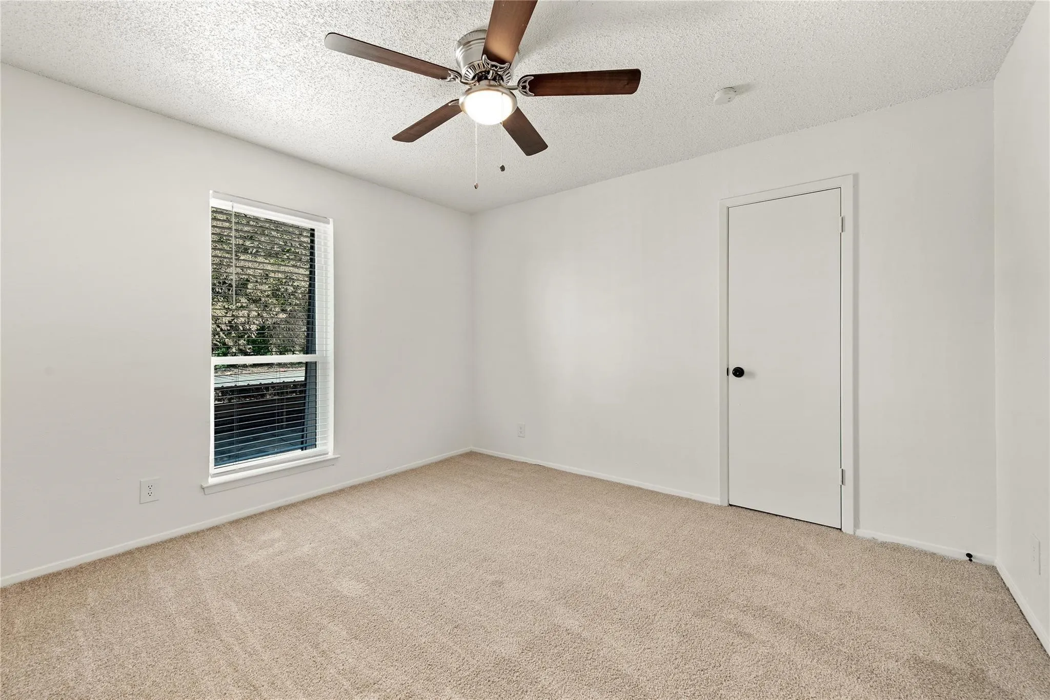 Neutral tone wall finishes, ceiling fan with integrated lighting, carpeted flooring, and a single window with blinds