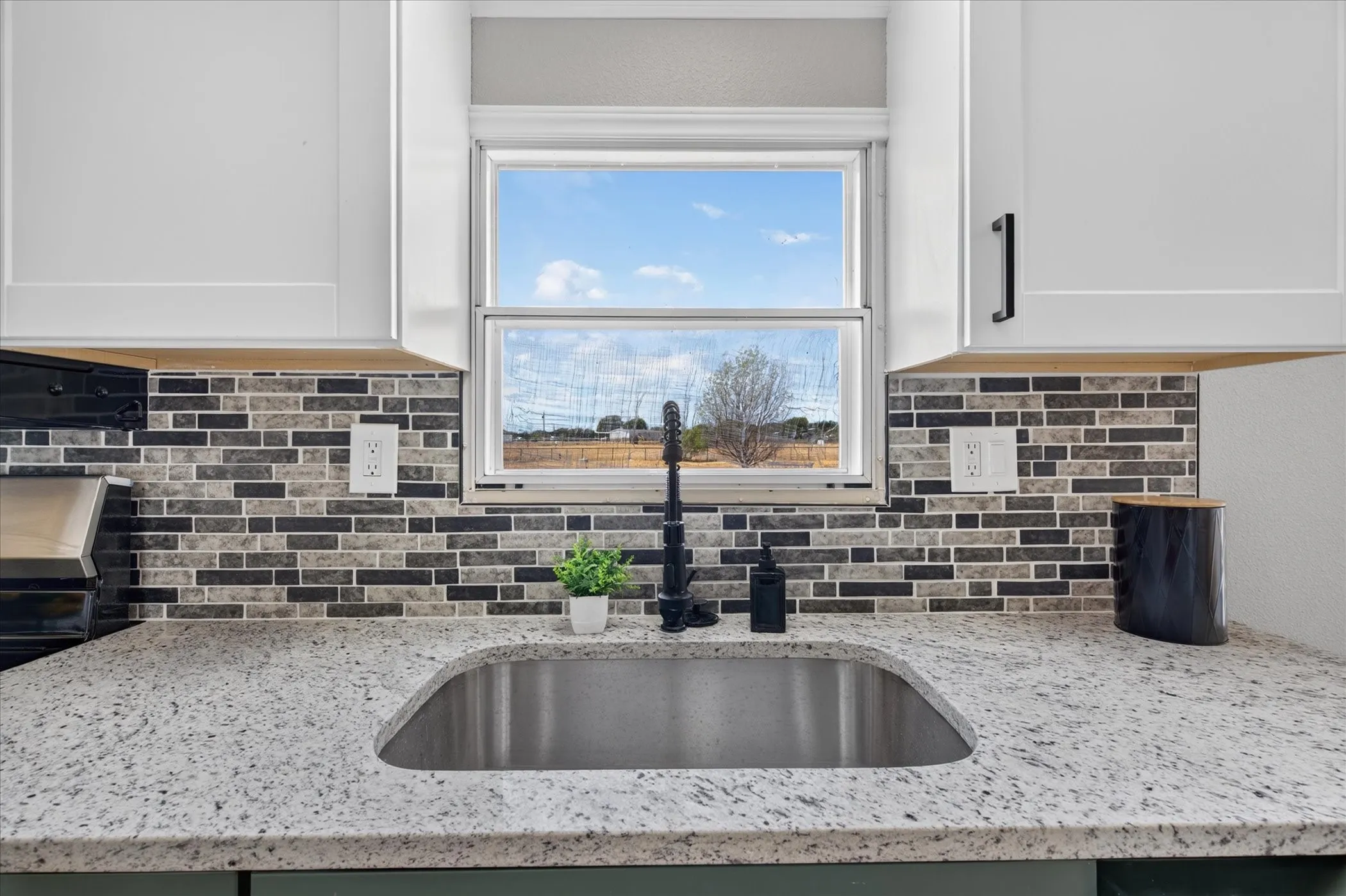 White shaker cabinetry, mosaic tile backsplash, stainless steel sink, black faucet, and speckled countertops
