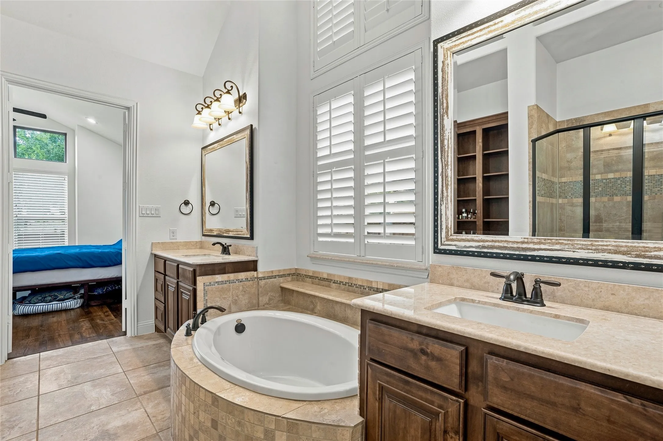 Dual vanities with undermount sinks, a built-in soaking tub with tile surround, plantation shutters, and tiled flooring