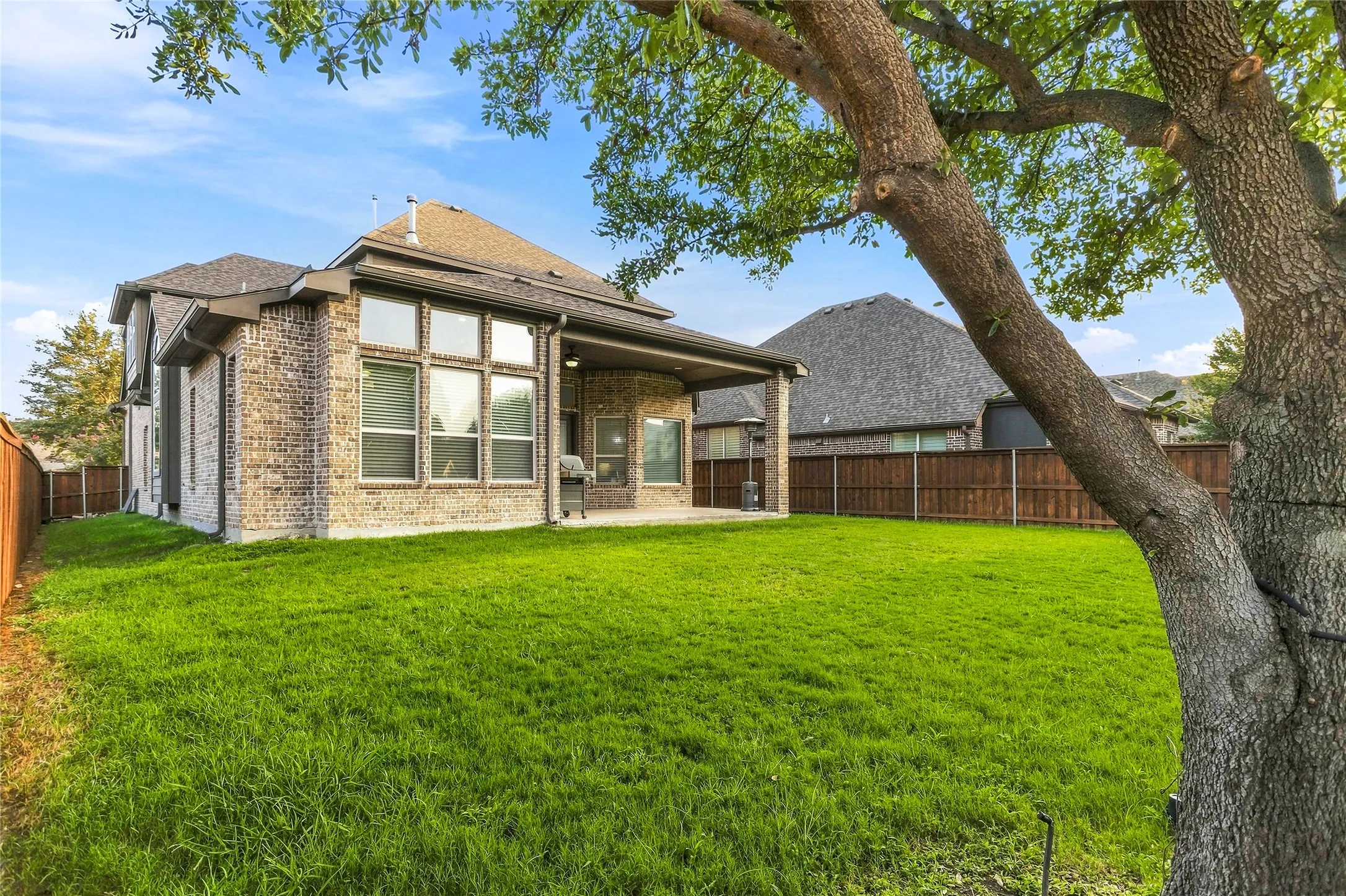 Expansive green lawn with a mature tree, featuring a brick exterior with multiple windows and a covered patio